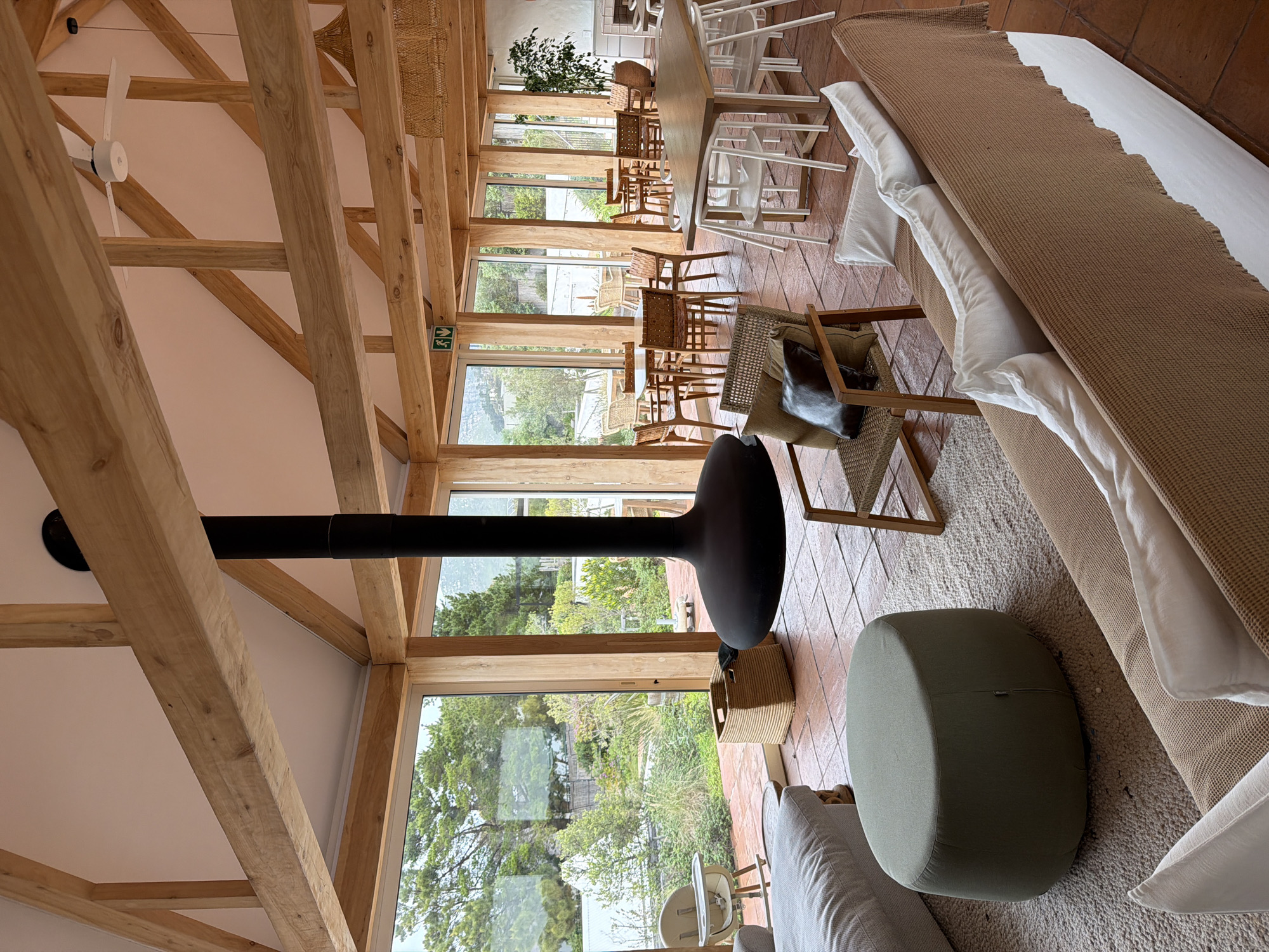 This image depicts the interior of a modern, open-plan restaurant or lounge area with a rustic design, featuring exposed wooden beams and large glass windows overlooking a lush, green outdoor space. The location is identified as Hout Bay, South Africa.
The focal point of the indoor space is a suspended, matte black, modern fireplace. The flooring is a terracotta tile. In the foreground, there is a light-colored, U-shaped sofa with white cushions and a brown textured throw draped over it. To the left, a large, round, olive-green ottoman sits on a cream-colored rug. Further left, partially visible, is another light-colored sofa.
The middle ground of the image shows several seating arrangements. To the left, there is a high chair. A woven basket is placed on the floor. In the center, a light-colored armchair with a woven seat and backrest is positioned, adorned with a dark brown cushion. Behind this armchair, a large dining table with white chairs is visible, with other smaller dining tables and chairs set up throughout the space.
The background reveals the expansive windows that offer a view of trees, foliage, and potentially a body of water or coastline, consistent with the Hout Bay environment. A ceiling fan is mounted on the wooden beam structure. In the far right background, part of a white wall and a fireplace mantel can be seen, along with a large potted plant. Two green directional signs with white arrows and figures are visible on the wooden pillar between window panes.
No people are present in the scene. The overall atmosphere is one of relaxed, contemporary, and nature-integrated dining or leisure.