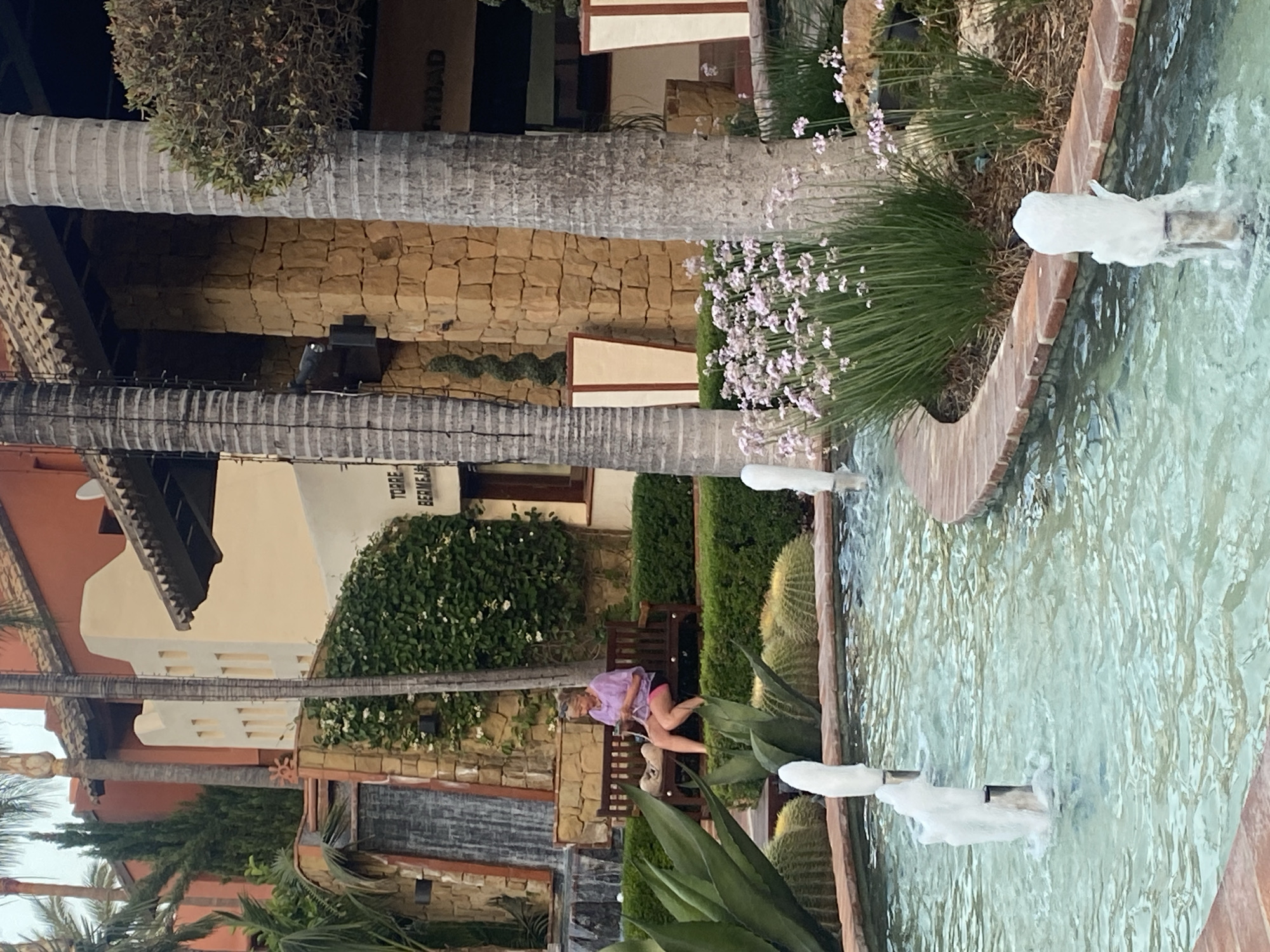 A woman sits on a wooden bench in a courtyard in Estepona, Spain. She is wearing a purple shirt and red shorts. The courtyard features a fountain with water cascading down, surrounded by lush green plants, including cacti and palm trees. The architecture of the buildings is visible, with stone walls and cream-colored facades. The time of day appears to be daytime, with ample natural light. The overall scene suggests a relaxed and tranquil atmosphere. Visible text on a building reads "TORRE BERMEJA".