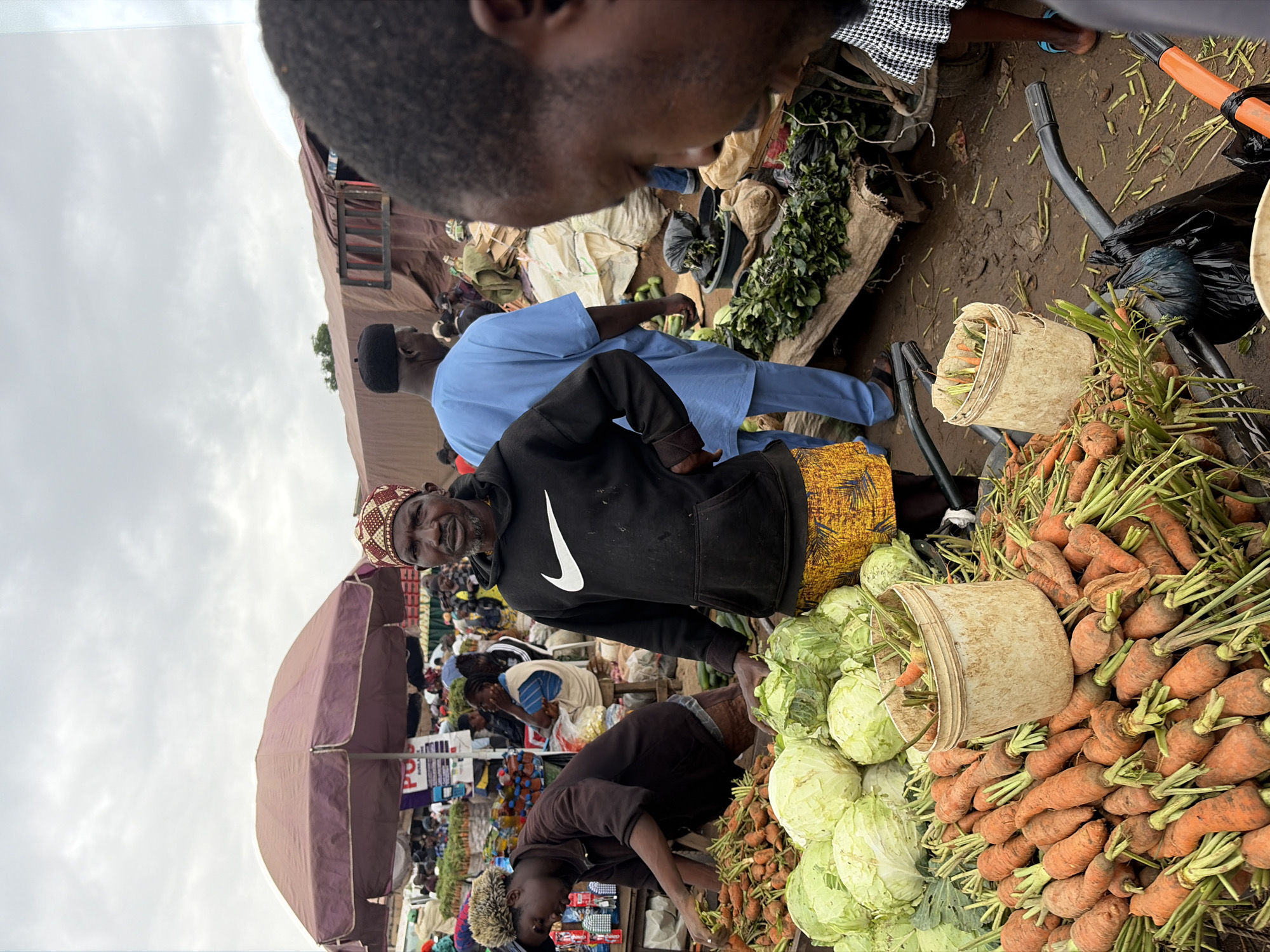 This image captures a bustling market scene in Gura Top, Nigeria, likely during daytime given the overcast sky. The photograph is oriented vertically. Several individuals are visible, engaged in commerce or interacting within the crowded environment.
In the foreground, a man wearing a black hoodie with a white Nike logo and a patterned cap looks towards the camera with a neutral expression. He is standing next to a large pile of fresh carrots, some of which are in a light-colored bucket. To his left, a man in a light blue traditional outfit is partially visible, facing away from the camera. Behind them, a dense crowd of people can be seen, with various goods and stalls suggesting a vibrant marketplace.
Further back, a portion of a large, light brown umbrella shades some of the stalls. The ground appears to be made of dirt, typical of an open-air market. The overall impression is one of a busy, active marketplace where goods are being sold. The weather appears to be cloudy, obscuring direct sunlight. There are no discernible timestamps or explicit text that would indicate the exact time of day or specific date.