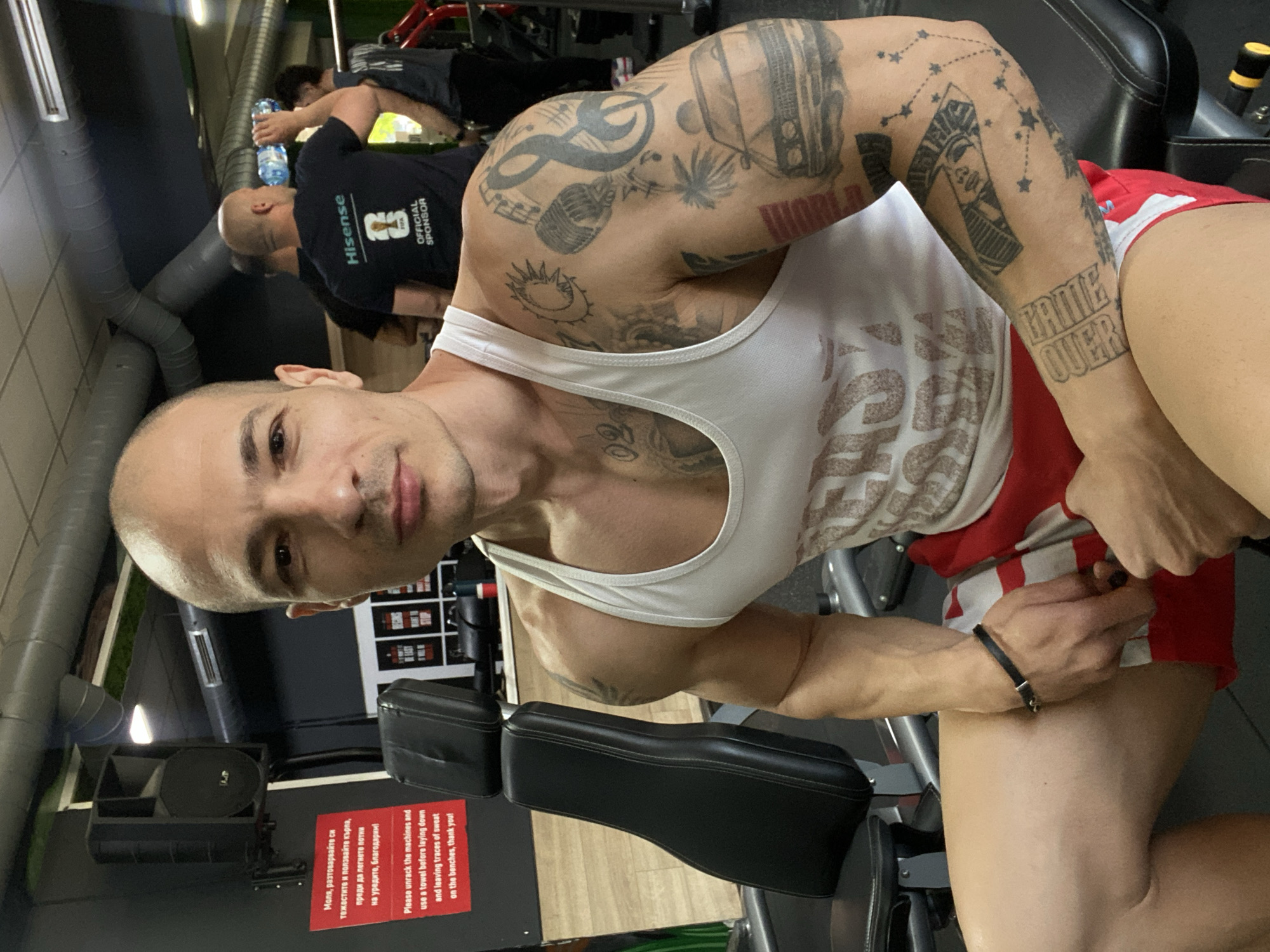 A man is pictured in a gym in Pleven, Bulgaria. He has a shaved head, a fair complexion, and various tattoos covering his arms and chest. He is wearing a white tank top and red and white striped athletic shorts. He is seated on a weight machine, looking directly at the camera.
In the background, another man is visible wearing a black t-shirt with the word "Hisense" and the words "Official Sponsor". He is bending over, potentially drinking from a water bottle. Gym equipment is visible around the two men.
There is a sign with red lettering on the wall that reads "Моля, разтоварвайте си тежестите и ползвайте кърпа, преди да легнете потии на уредите, благодарим! Please unrack the machines and use a towel before laying down and leaving traces of sweat on the benches, thank you!"