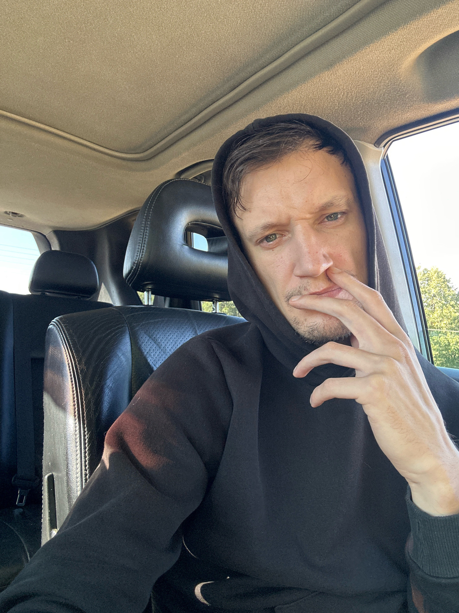 A man in a black hoodie sits in the passenger seat of a car, his hand touching his nose and mouth, with a thoughtful expression. The interior of the car, including leather seats and a sunroof, is visible. Outside the car, trees and a clear sky suggest an outdoor setting. The photo appears to have been taken during daylight hours, with sunlight casting shadows on the car's interior and the man's face. There is no information available that places this image specifically in the city of Makiv, Ukraine.