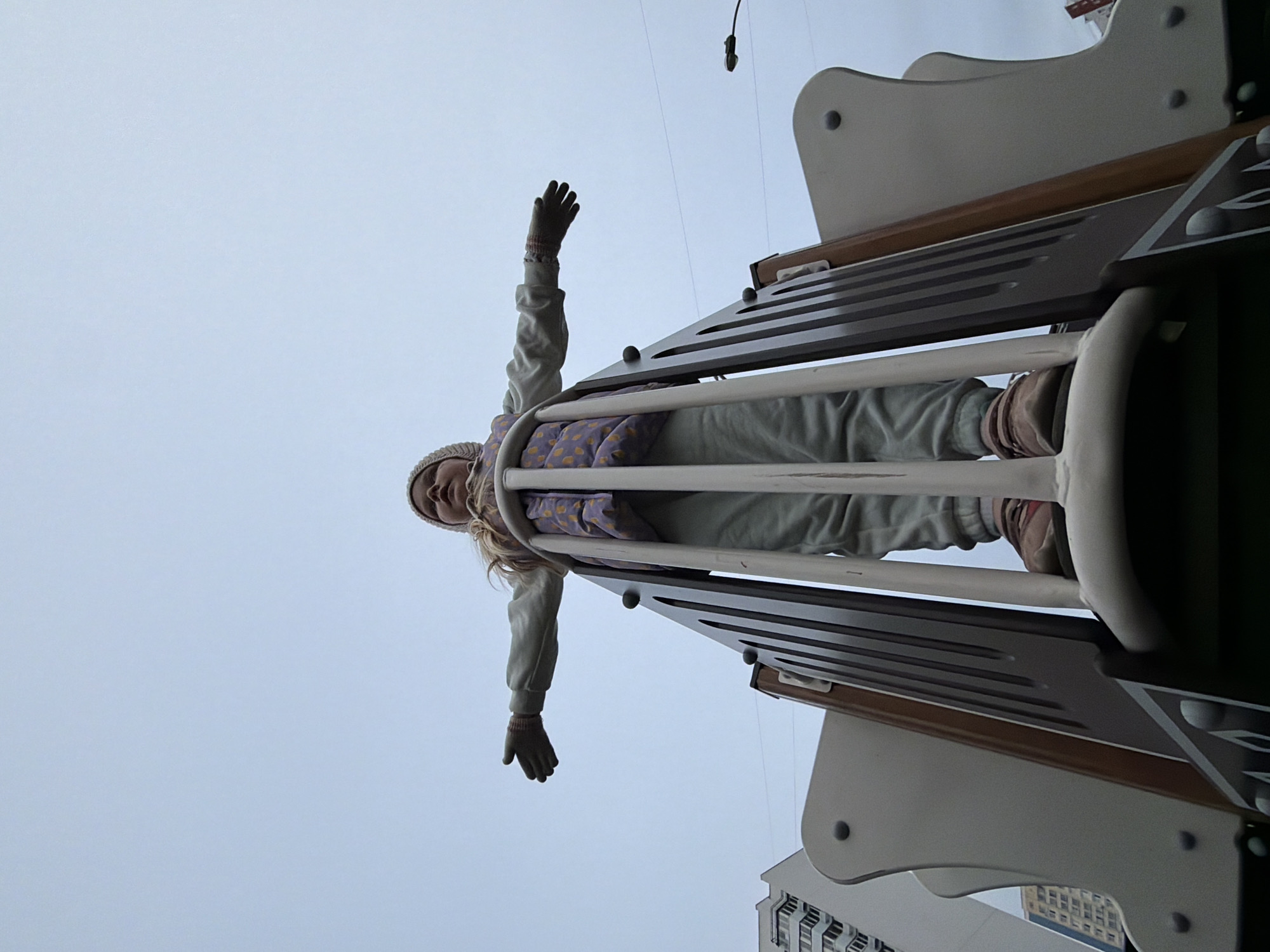 A young child, likely a girl, is captured from a low-angle perspective, positioned on what appears to be a playground slide or climbing structure. She is wearing warm clothing, including a light-colored knit hat, a long-sleeved top, and gloves, suggesting cool or cold weather. Her arms are outstretched to her sides, conveying a sense of playfulness or "flying" as she looks down towards the camera with a subtle expression.
The playground structure features grey and dark brown elements, with horizontal slats and light-colored railings. The background consists almost entirely of a vast, overcast grey sky, indicating diffused lighting and potentially a dull day. Faint outlines of urban buildings are visible in the distance at the bottom right, suggesting an urban or suburban setting. The overall scene captures a moment of childhood joy and freedom on a playground in Moscow, Russia. No visible text is present in the image.