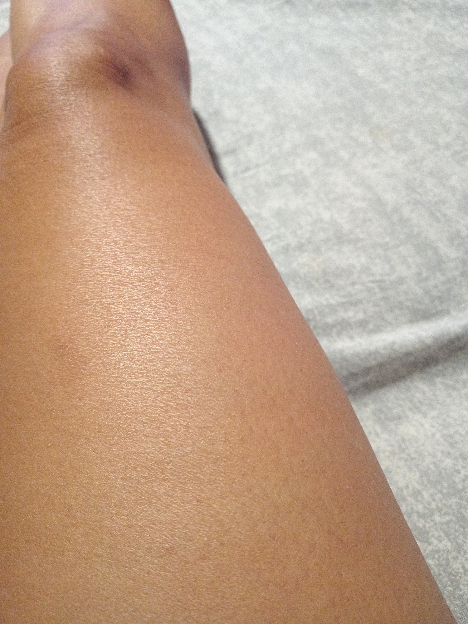 The image displays a close-up view of a person's leg, with the skin appearing smooth and lightly tanned. The leg is positioned on a gray, textured surface, possibly a blanket or bedsheet, which is slightly wrinkled. The lighting suggests an indoor environment. There is no discernible urban or environmental context to associate this image with Novopokrovka, Russia, or any other specific location. The image focuses solely on the texture and coloration of the skin and the immediate surface beneath it.