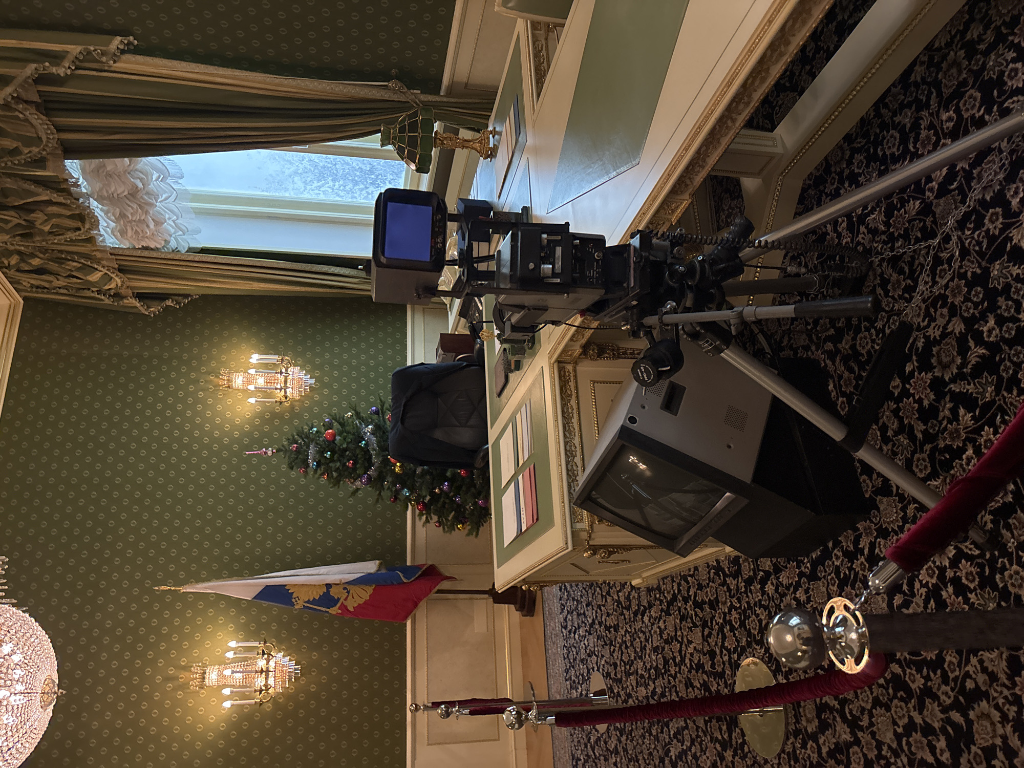 This detailed image depicts a formal, ornate room, possibly an office or study, set up for a video broadcast or recording in Yekaterinburg, Russia. The room features dark green walls with a subtle repeating pattern, accented by off-white wainscoting with gold trim. A large crystal chandelier hangs from the ceiling, and two ornate gold wall sconces with candle-like bulbs illuminate the left wall. The floor is covered by a dark carpet with a light-colored floral or scroll pattern. A tall, decorative window on the right side of the room is adorned with layered green and gold curtains, and a sheer inner curtain; the view outside suggests a cold, possibly snowy, environment.
In the center, a large, ornate executive-style desk with cream-colored panels, gold accents, and green desktop inserts is positioned. A black, padded office chair is visible behind the desk. To the left of the desk stands a Russian Federation flag on a dark wooden base. To the right, a decorated Christmas tree with colorful spherical ornaments and a finial top adds a festive element.
Broadcasting equipment is prominently featured in the foreground: a professional video camera, labeled with 'Canon FPD-40' on its lens control, is mounted on a tripod and points towards the desk. A small monitor attached to the camera displays a blue screen. On the floor, to the left of the camera setup, a larger, older-model CRT monitor is placed, showing a dark screen. On the desk, several stacks of papers or booklets with colored covers (red, white, blue, green, yellow, orange) are neatly arranged on the left side, and a green-shaded banker's lamp with a gold base is on the right. In the lower left foreground, two chrome stanchions connected by a red velvet rope cordon off an area. No individuals are visible in the scene.
