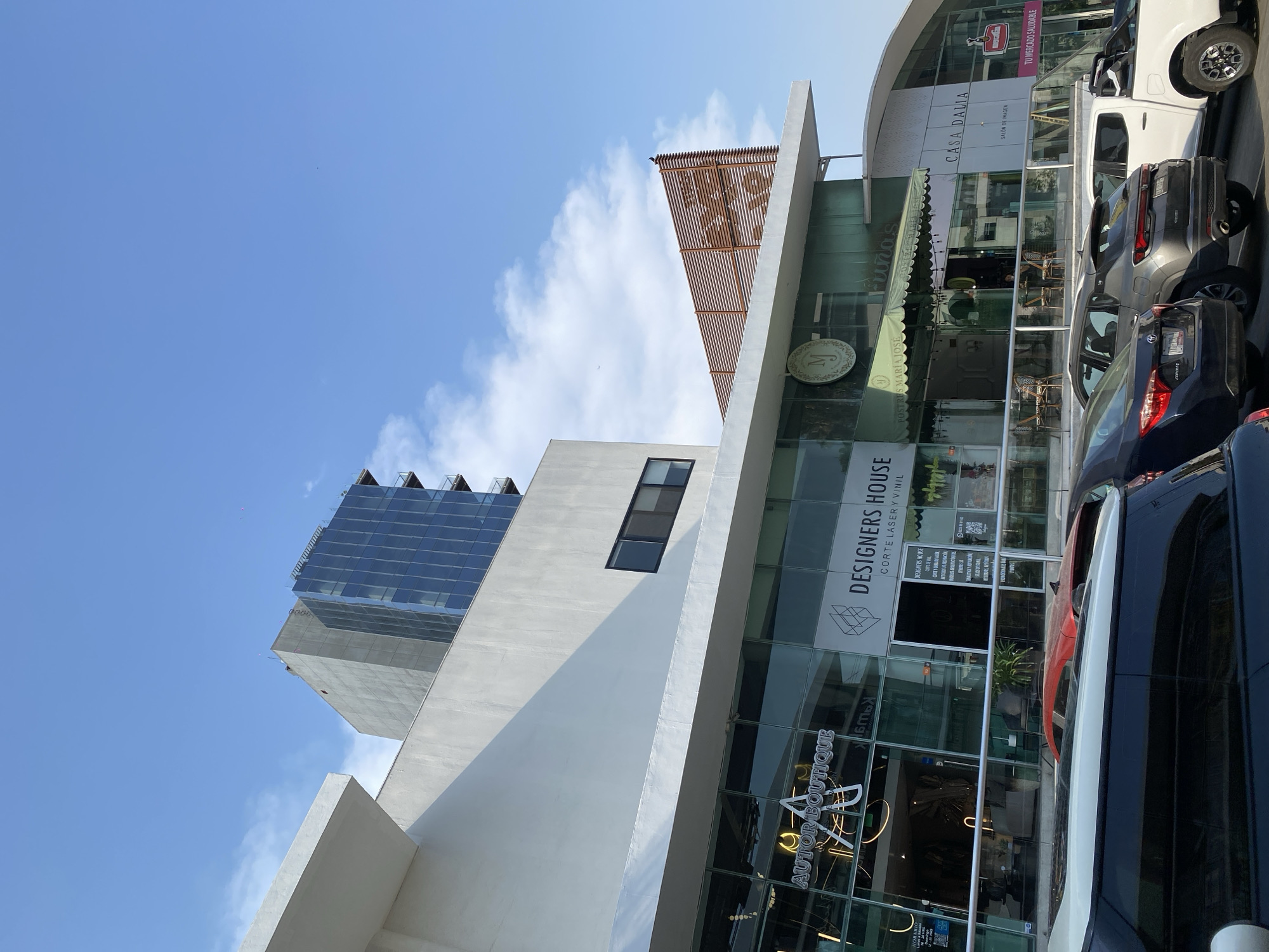 The image is a low-angle, exterior shot of a commercial building complex in an urban setting. The sky is partly cloudy with a clear blue hue. The building features modern architectural design with glass and white concrete as primary materials. Various businesses are visible including "Autor Boutique," "Designers House" offering laser cutting and vinyl services, "Casa Dalia Salon de Imagen" and what appears to be a shop named "Postres Maria Jose," recognizable by a stylized “M” logo. Several parked cars partially obstruct the view of the storefronts, indicating an active commercial area. In the background, a skyscraper under construction can be seen with an unfinished framework, suggesting ongoing urban development. The geographical location is unknown as no identifying details like street names or well-known landmarks are visible.