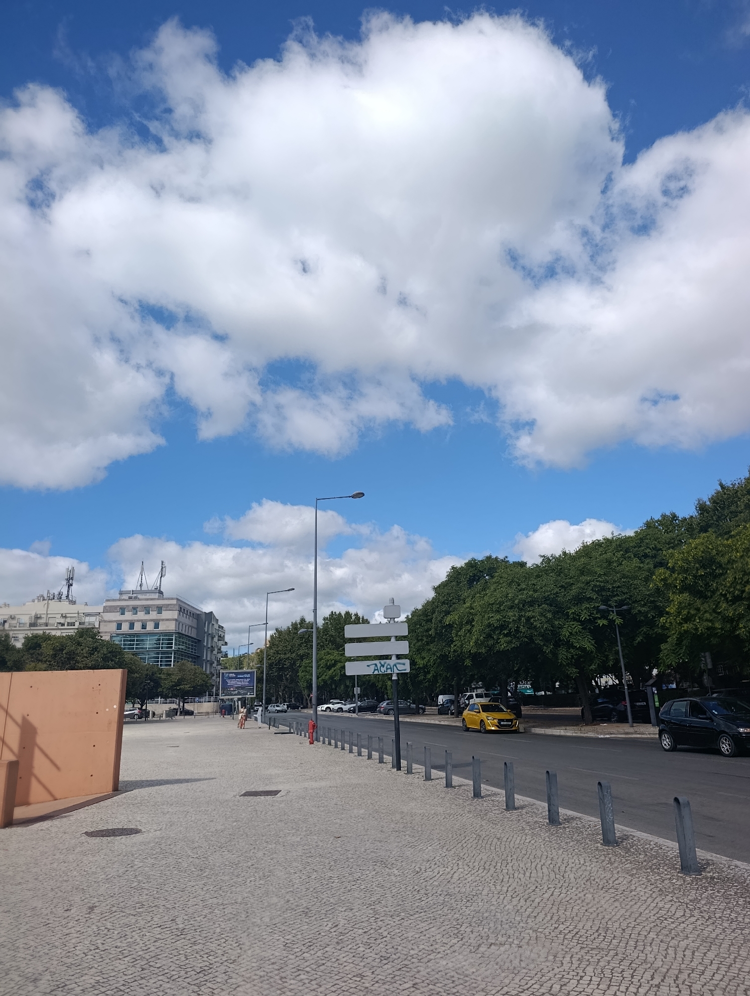 The image depicts a street scene in Setúbal, Portugal, under a bright blue sky with scattered white clouds. In the foreground, a cobblestone pavement leads to a road lined with metal bollards. Across the road, dense green trees form a natural barrier. Beyond the trees, a modern building with glass windows and balconies is visible, with smaller buildings and communication towers in the background. Several cars are parked or driving on the road, including a bright yellow vehicle. A person is walking in the distance on the pavement. The scene suggests a pleasant day in an urban environment.