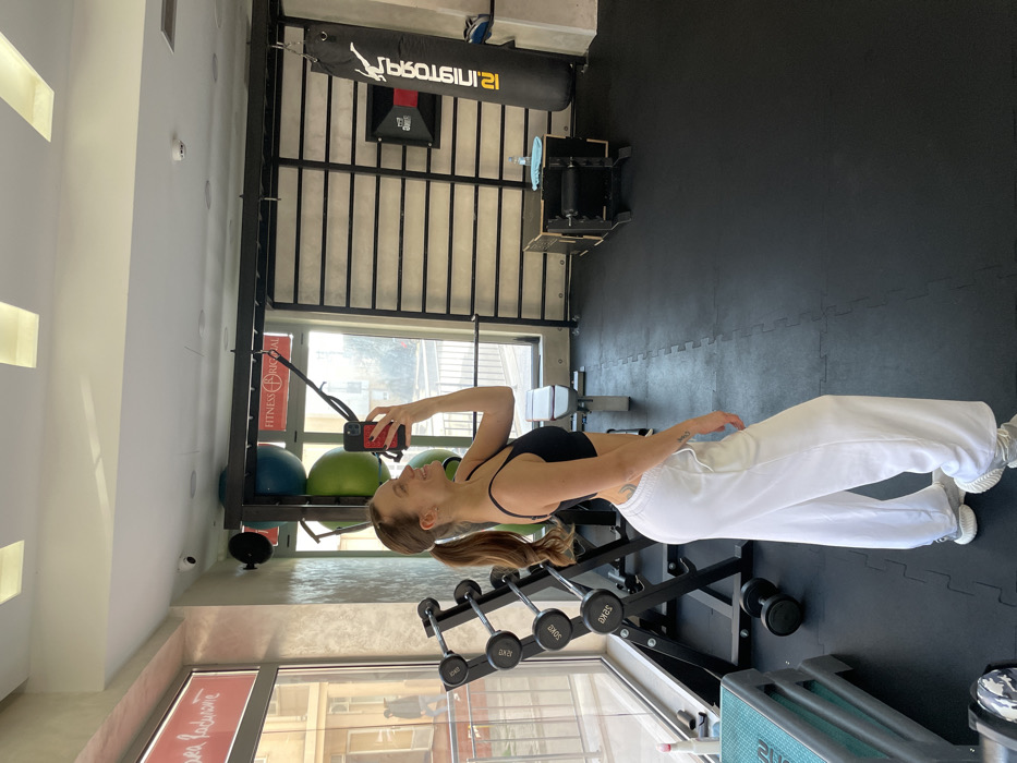 The image depicts an interior scene within a fitness gym identified as Fitness Original, located in Budva, Montenegro. The primary subject is a female positioned in the center-left, observed in a slightly turned profile view, facing towards the left. She is wearing a black sports bra, white wide-leg sweatpants, and light grey athletic shoes. Her brown hair is pulled back into a ponytail. She holds a red-cased smartphone in her raised right hand, capturing a mirror selfie. A small tattoo is visible on her left hip area.
The gym floor is uniformly covered with black interlocking rubber mats. To the left, large windows provide an exterior view of urban buildings and a street. Adjacent to the window, a black metal rack holds multiple pairs of dumbbells of varying weights. A light blue step platform is partially visible in the lower-left foreground.
In the background, a black metal functional training rig is visible, featuring horizontal wall bars. Above the rig, two large green exercise balls are stored on a shelf. A black punching bag, marked with white Cyrillic text "PROTOTIP", hangs from the rig. Beneath the punching bag, a dark wooden plyometric box is present, with a light blue towel draped across its top surface.
Exterior signage visible through the windows includes a red sign bearing the text "FITNESS ORIGINAL" and another partially obscured red sign with Cyrillic characters. The ceiling is white, incorporating rectangular recessed lighting panels. A security camera is mounted on the ceiling in the upper-left, and a smoke detector is located on the ceiling in the upper-right.