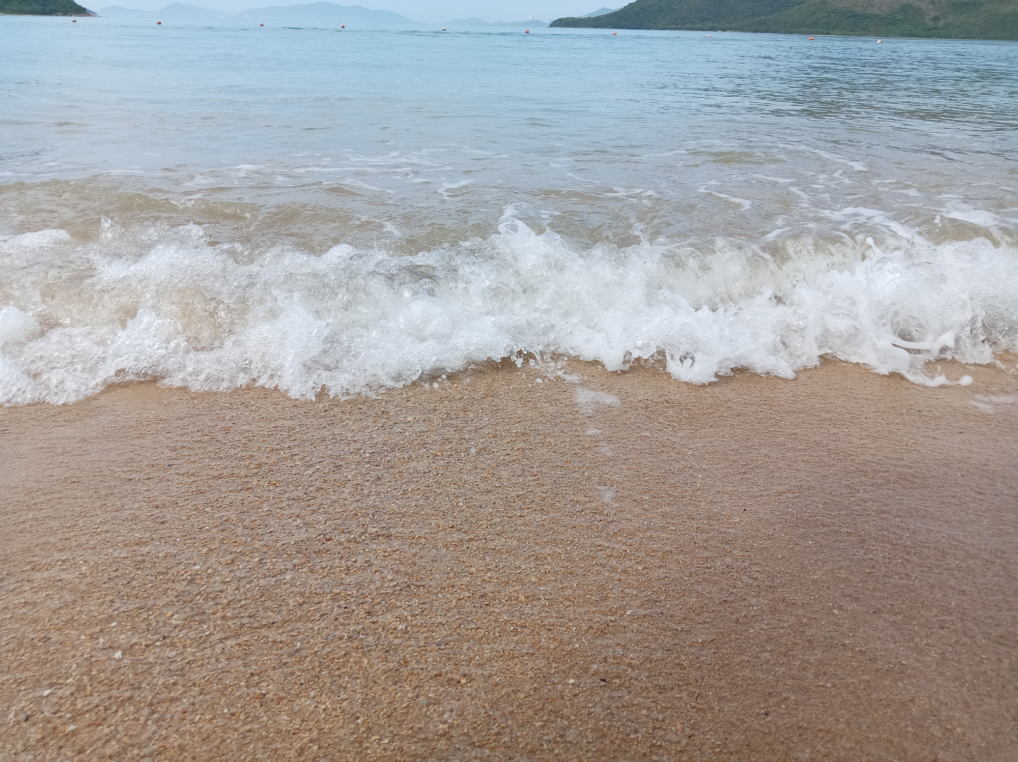 The image depicts a serene beach scene in Hoa Thang, Vietnam. A gentle wave is seen lapping at the sandy shore, its white foam creating a delicate line between the water and the beach. The water is a soft blue, merging into a lighter hue towards the horizon where distant hills are visible under a hazy sky. Several small, orange buoys are scattered in the calm sea, marking a designated area or guiding vessels. The sand is fine-grained and light brown, appearing wet and smooth where the water has receded. No people are visible in the frame. The overall atmosphere is tranquil and natural.