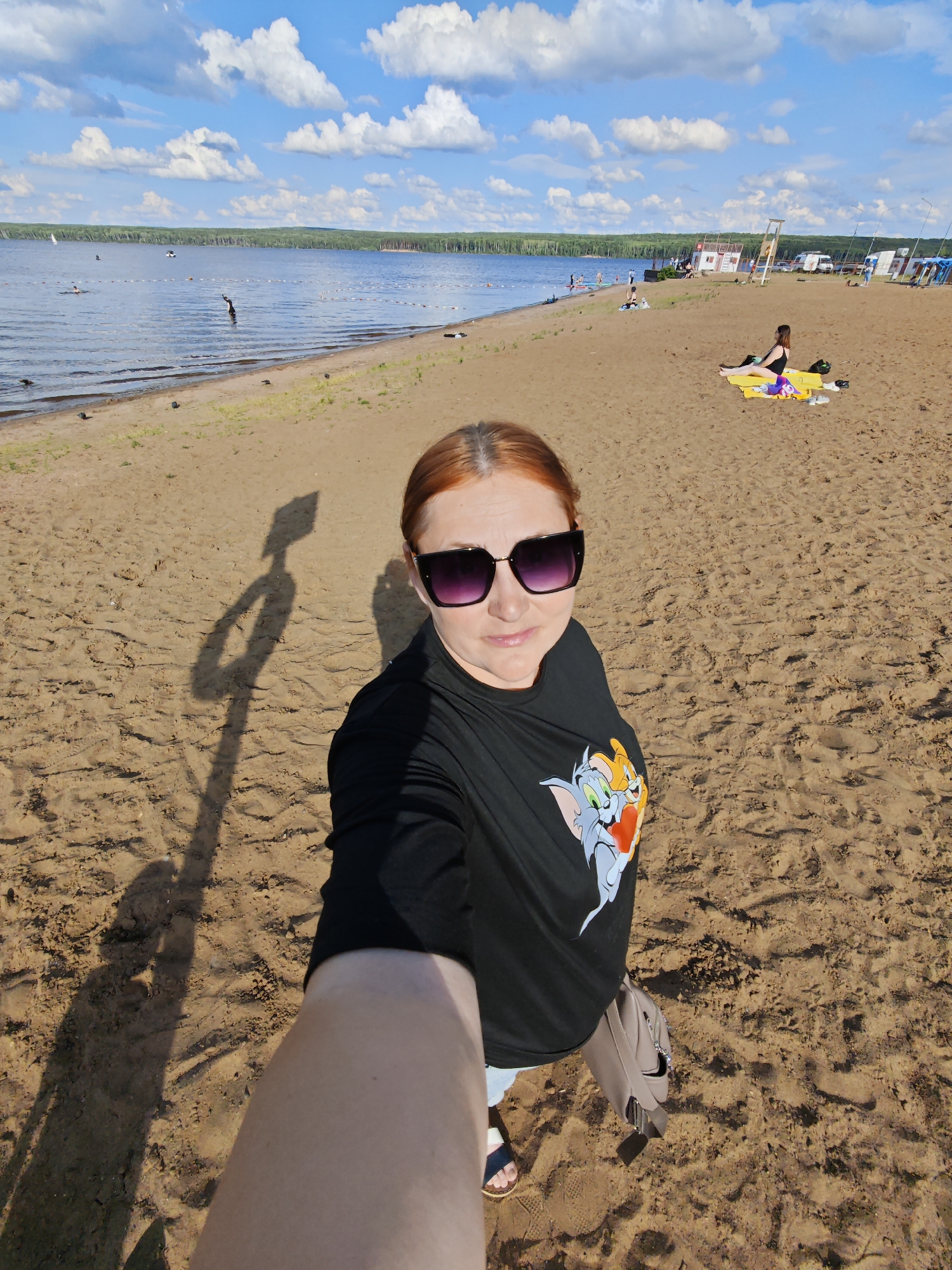 A woman with auburn hair and sunglasses takes a selfie on a sandy beach. She is wearing a black t-shirt with a Tom and Jerry graphic. Her shadow is cast on the sand, alongside the shadow of a lamppost or similar pole. In the background, the water of a lake is visible, with people swimming and wading. Further back, a tree line borders the lake, and a few buildings and vehicles are present on the shore. The sky is blue with scattered white clouds. The scene appears to be the beach at the Kama River in Chaikovsky, Russia.