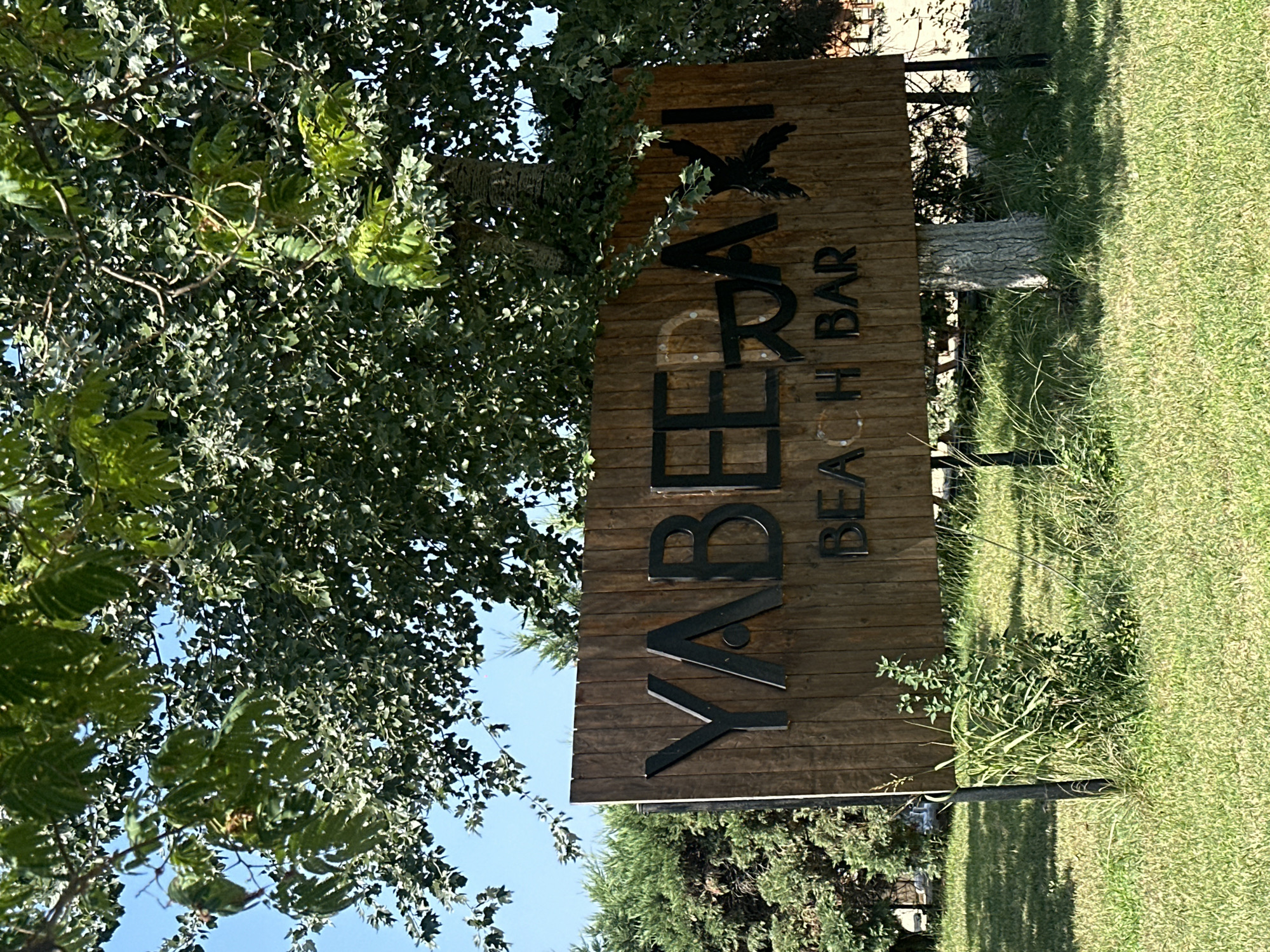 A wooden sign with the words "YABERAXI BEACH BAR" is prominently displayed in front of lush green foliage and grass under a clear blue sky. The sign has large, dark, raised letters spelling out "YABERAXI" and smaller, raised letters spelling out "BEACH BAR" below it. The sign is supported by dark metal posts. The scene appears to be outdoors, likely in a park or recreational area. The location is Ofrynio Beach, Greece.
