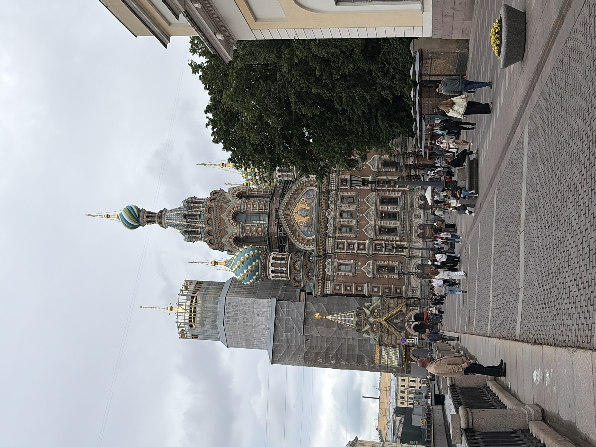 The image shows a view of the Church of the Savior on Spilled Blood in Saint Petersburg, Russia. The church, known for its ornate Russian Revival architecture and vibrant mosaic domes, is partially covered in scaffolding, suggesting ongoing renovation or construction work. One of the iconic onion domes with blue and green patterns is visible. In the foreground, a paved walkway runs alongside a granite railing. Several people are walking on the walkway and standing in front of the church, some holding umbrellas, indicating it might be a cloudy or drizzly day. The sky is overcast, typical of Saint Petersburg's weather. Behind the church, a large tree provides a lush green contrast to the architecture. The scene appears to be a typical day in a tourist area, with people observing the landmark. No visible text is present in the image.