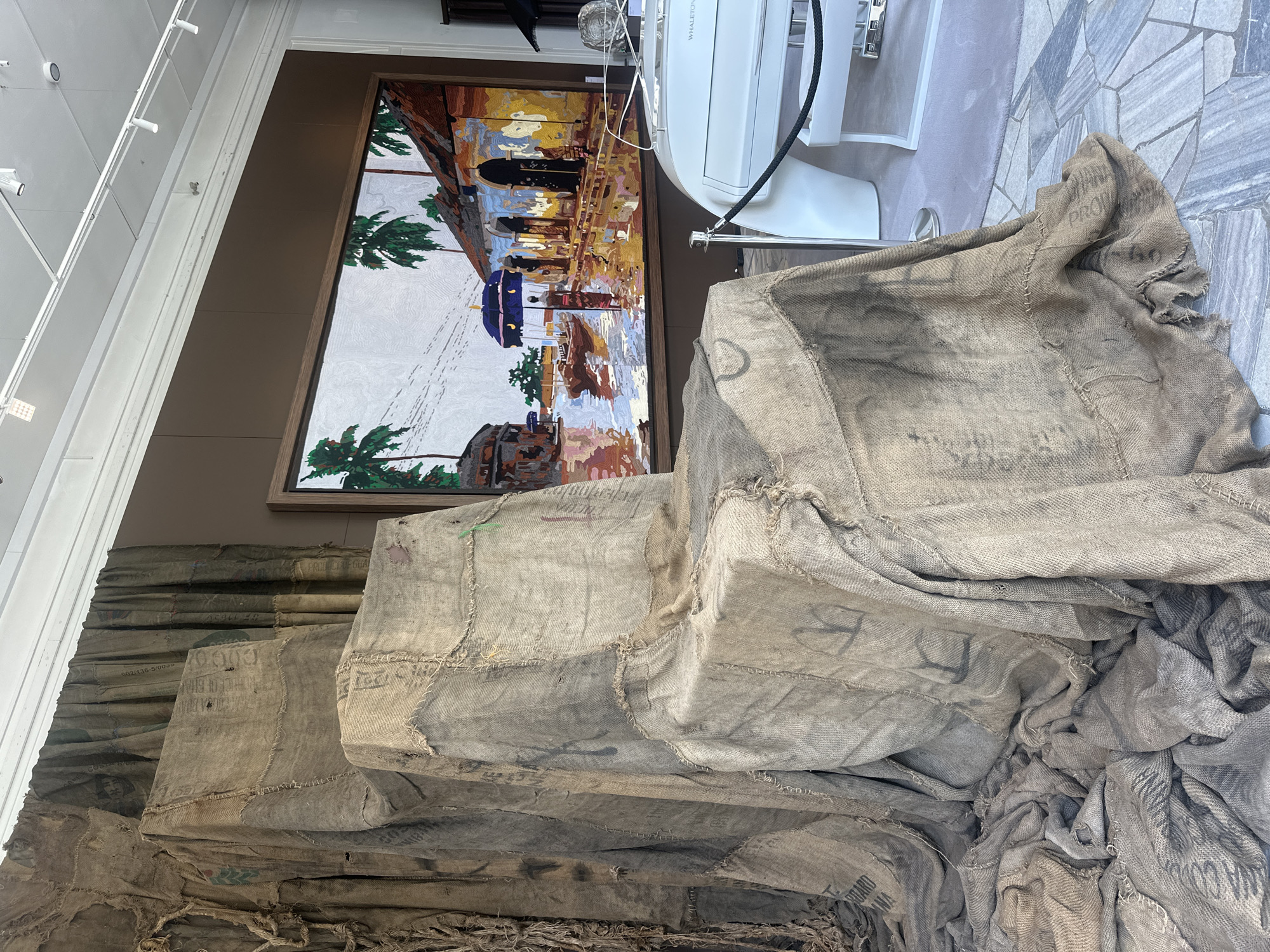 A still life photograph taken from a medium distance showcases an art installation featuring a large, throne-like structure draped in burlap fabric, positioned on a patterned tiled floor. The burlap, sewn together in patches and showing signs of wear, creates folds and textures that suggest a sense of aged materiality. Behind the burlap structure, a colorful, framed painting depicts a waterfront scene with buildings, palm trees, and figures under umbrellas, rendered in a mosaic or tapestry style. To the right of the installation, a portion of a white boat or yacht console is visible, with a black rope cordoning off the area. The background wall is a uniform brown, and the ceiling features a track lighting system with adjustable spotlights. The scene is well-lit, with the lighting casting subtle shadows and highlighting the textures of the materials. The location is identified as the interior of a gallery or exhibition space. No individuals are present within the frame, and no specific action is depicted. The overall impression is one of curated display, emphasizing texture, color, and artistic presentation.