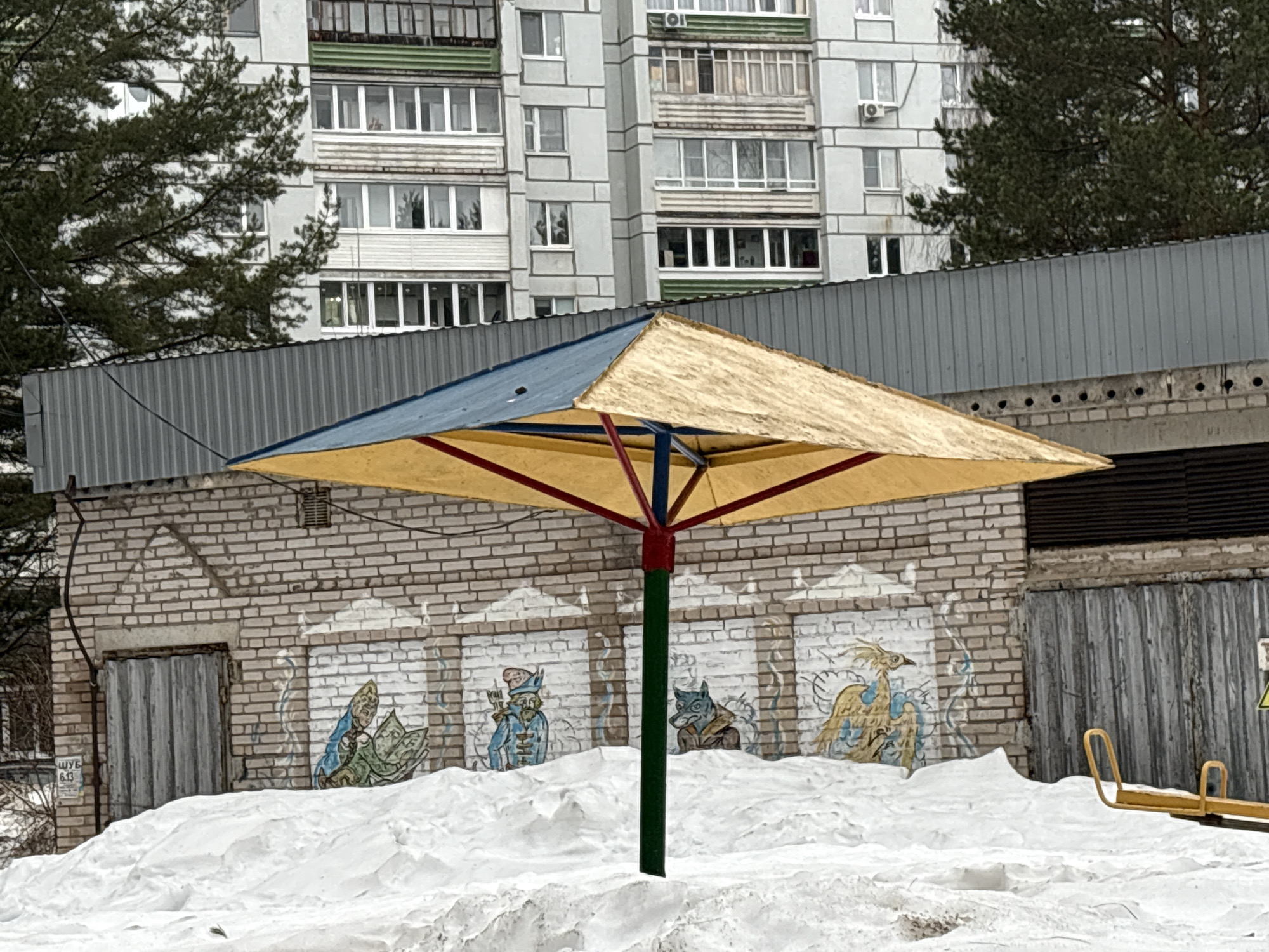 The image depicts a playground carousel in Chaikovsky, Russia, during winter, partly buried in snow. The carousel has a square, faded blue and yellow canopy supported by red struts and a green central pole. Behind the carousel is a brick wall with naive paintings of fairytale characters. A metal fence and an apartment building are visible in the background. The scene appears deserted and cold.