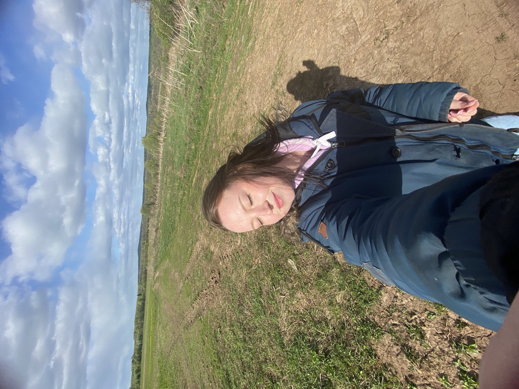 A woman is lying on a grassy field under a sky filled with white clouds. She is wearing a blue jacket and a pink hoodie. Her eyes are closed. The shadow of a person is visible near her. The background shows a field and some trees. The location is presumed to be in Дедушкино, Russia.