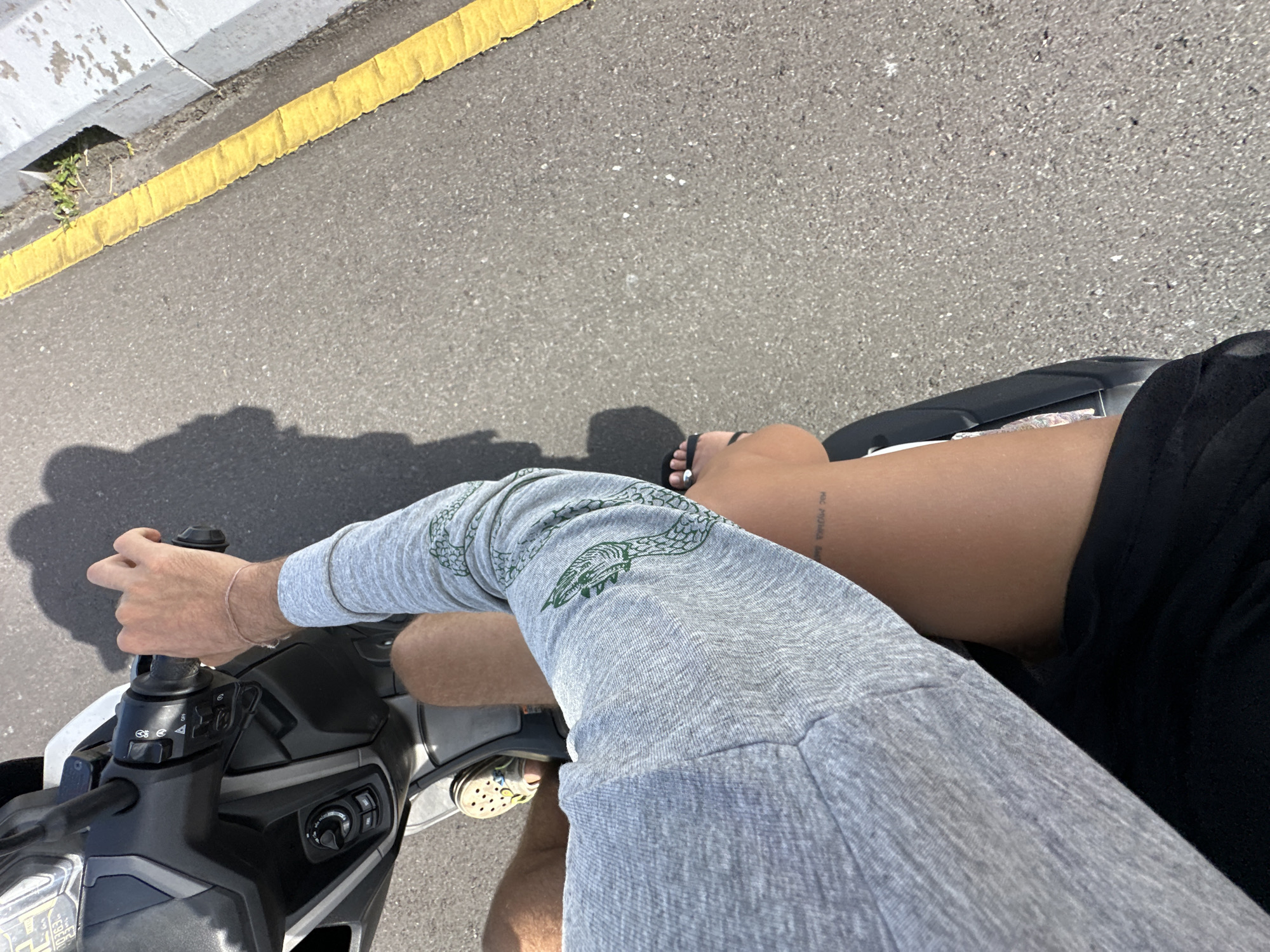 A person is riding a scooter on an asphalt road in South Denpasar, Indonesia. The road has a yellow curb. The person is wearing grey sweatpants with a reptile design, a black shirt, and Crocs. Their skin is tanned, and they have a tattoo on their leg. They are also wearing a bracelet. The scooter is black and white. The weather appears to be sunny, casting a shadow to the left of the scooter.