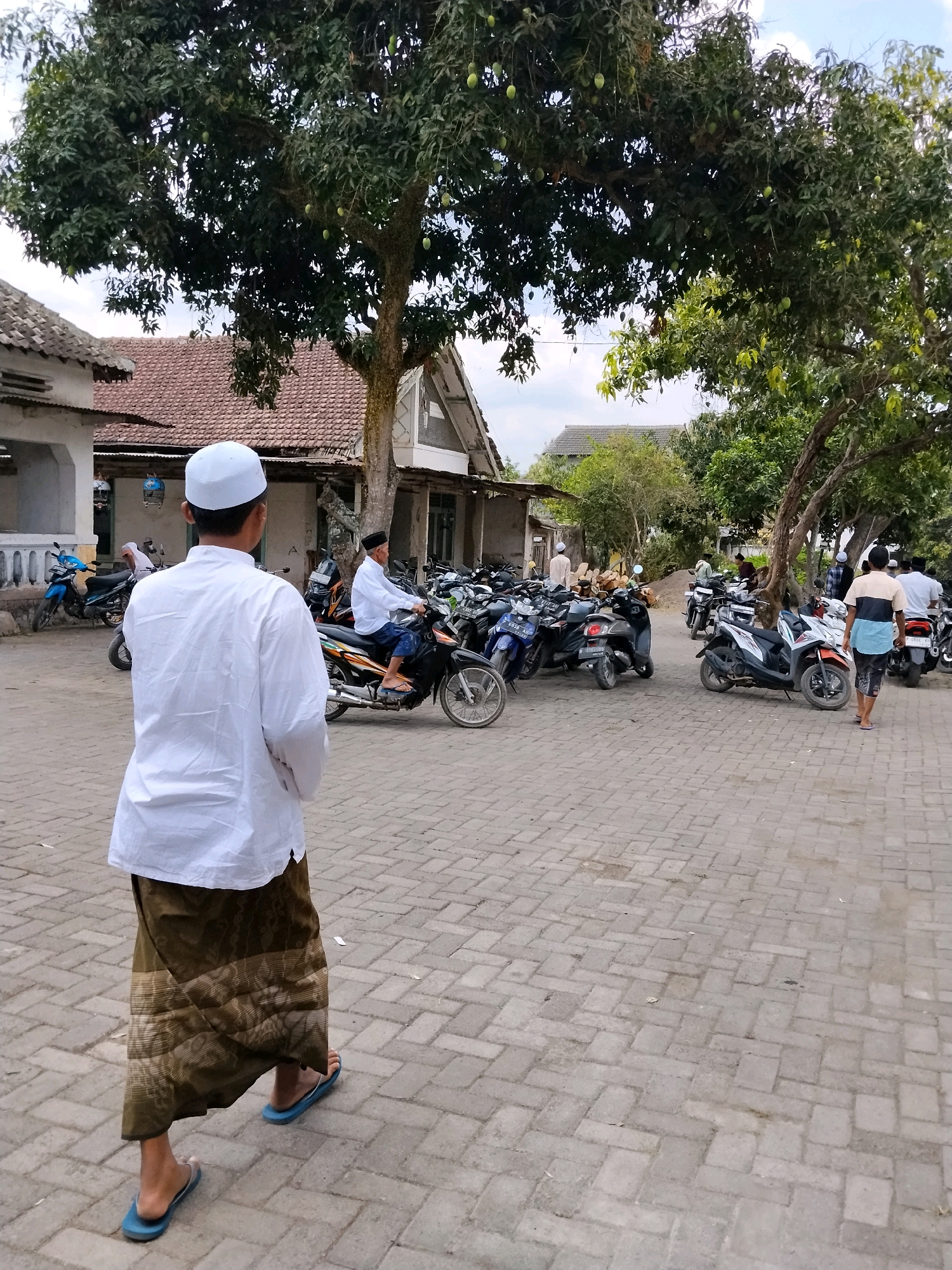 In Tlogosari, Indonesia, a young man wearing a white kufi and a white shirt with a patterned sarong walks away from the camera on a paved courtyard. Several motorcycles are parked in the background, and a few people are seen interacting with them. A large tree with green mangoes grows in the center of the frame, casting shadows on the scene. Buildings with terracotta roofs are visible behind the tree and motorcycles. The sky is overcast.
