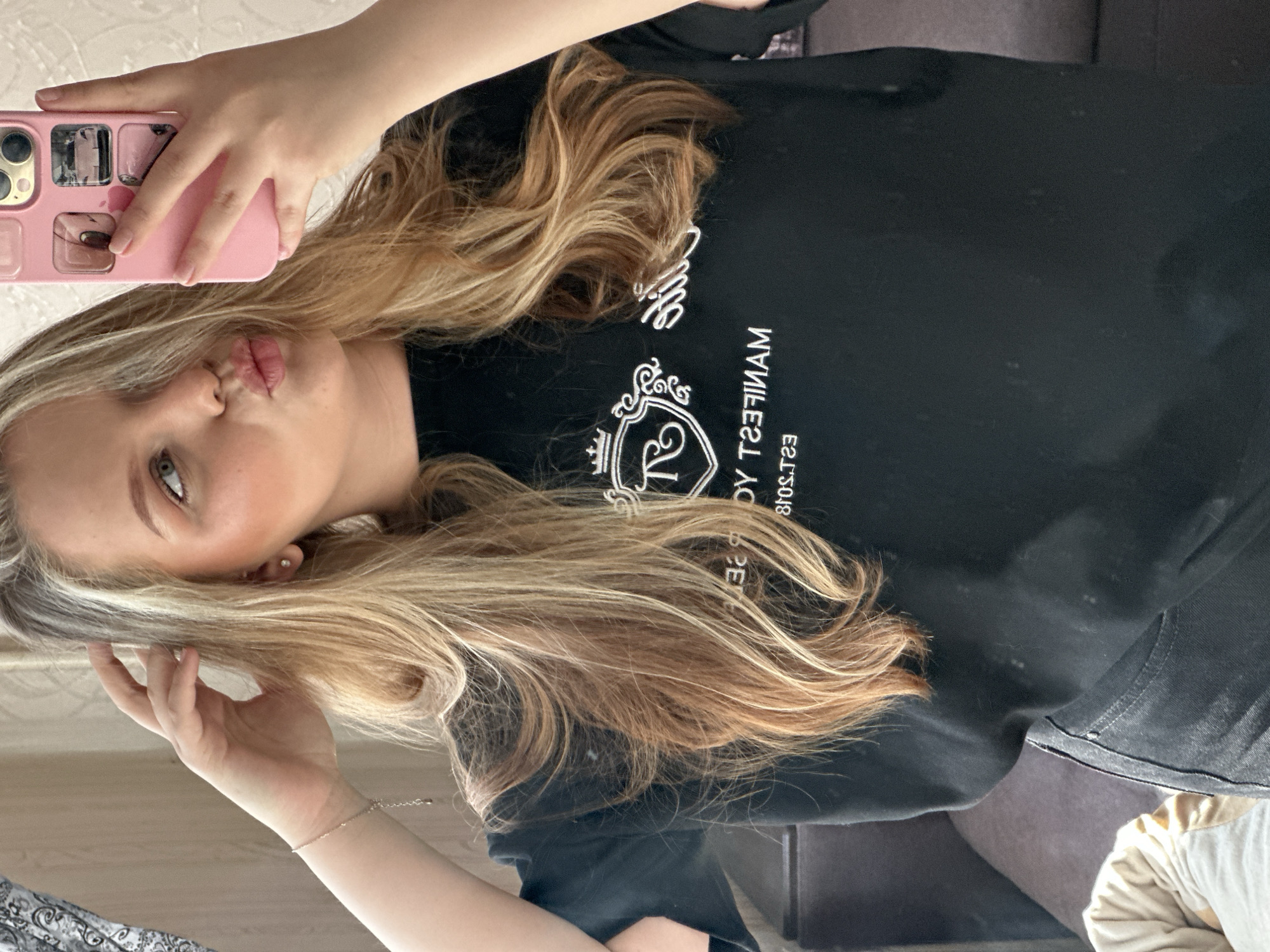 A woman with long, wavy blonde hair takes a selfie using a pink phone case. She is pouting her lips. She wears a black shirt with white writing. The location is not identifiable as Noviy, Russia from the provided image.
