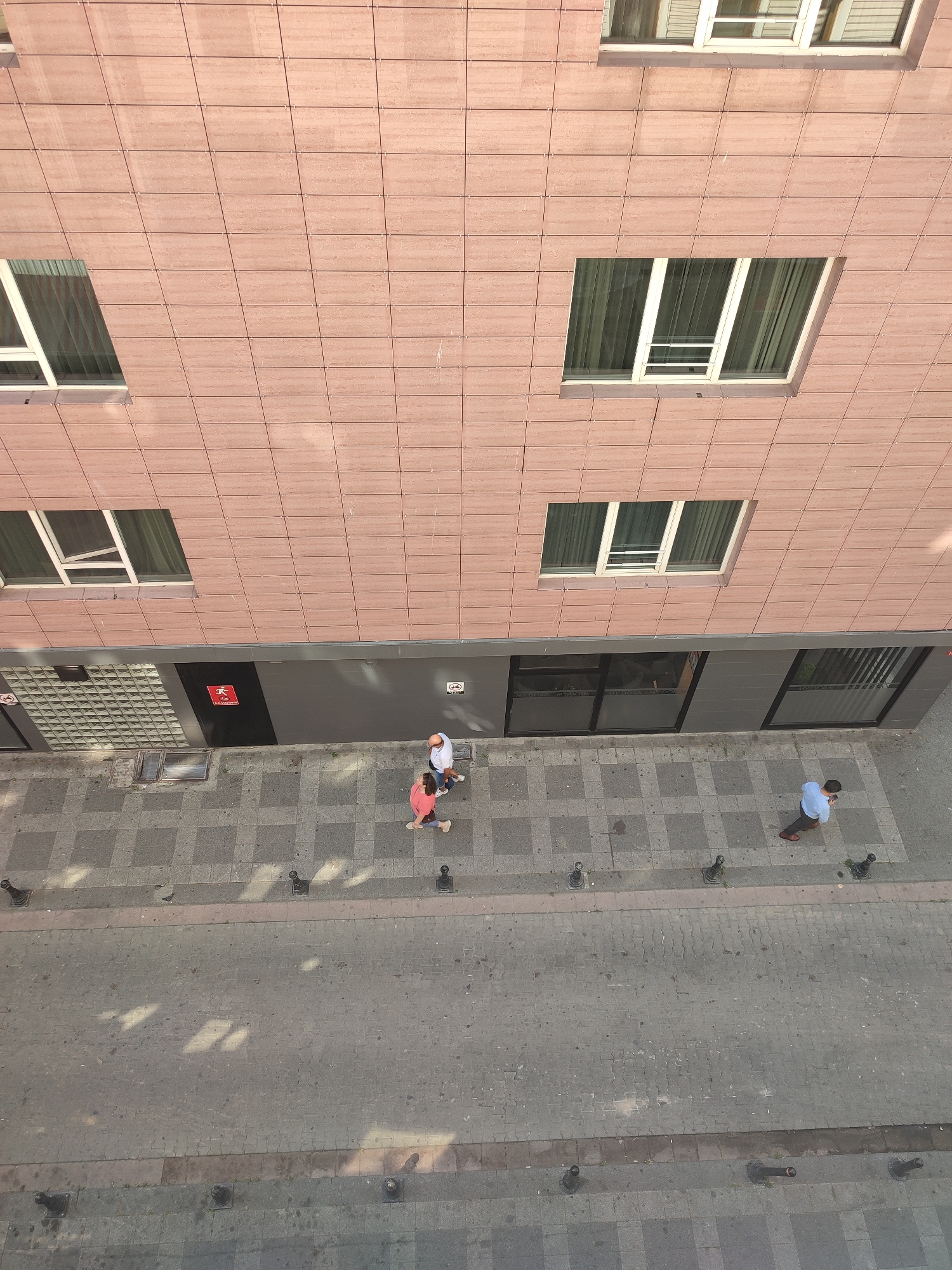 This is an overhead, high-angle shot of a street scene in Kadıköy, Turkey. The facade of a building with pinkish-brown tiled siding dominates the upper portion of the image, featuring multiple rectangular windows with white frames and sheer curtains. Below the building facade is a dark gray band, beneath which are store windows, one with frosted glass and black lettering that appears to read "MİROSA" multiple times.
On the sidewalk adjacent to the building, a man wearing a light blue shirt and brown pants is walking away from the camera, looking down at his phone. Further down the sidewalk, a couple is walking towards the camera. The woman is wearing a coral-pink shirt and light-colored pants, while the man is wearing a white t-shirt and dark shorts. They are walking side-by-side, with the man appearing to be talking.
The sidewalk is paved with a pattern of gray square tiles, separated by a narrower strip of textured pavement. A street runs alongside the sidewalk, made of gray interlocking pavers. Several black bollards are positioned along the edge of the sidewalk, separating it from the street. Sunlight casts shadows on the pavement, indicating a sunny day.
