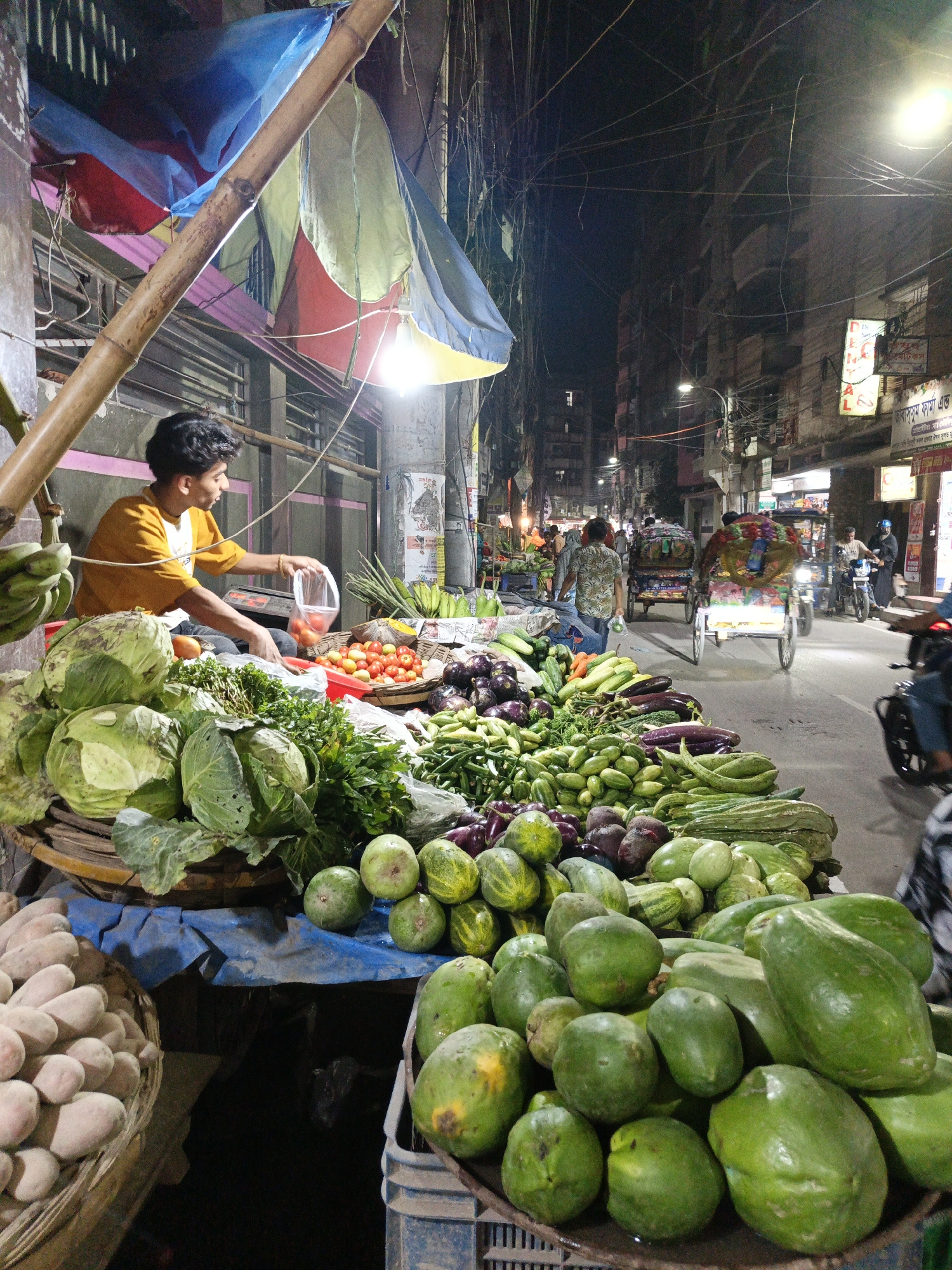 A vibrant street market in Dhaka, Bangladesh at night features a young man in a yellow shirt attending a vegetable stall. He is packaging tomatoes. The stall is laden with various produce including cabbages, greens, tomatoes, eggplants, cucumbers, and green papayas. The scene includes a narrow street with people, bicycle rickshaws, and motorcycles navigating the dimly lit road, with buildings towering overhead amidst a tangle of overhead wires.
