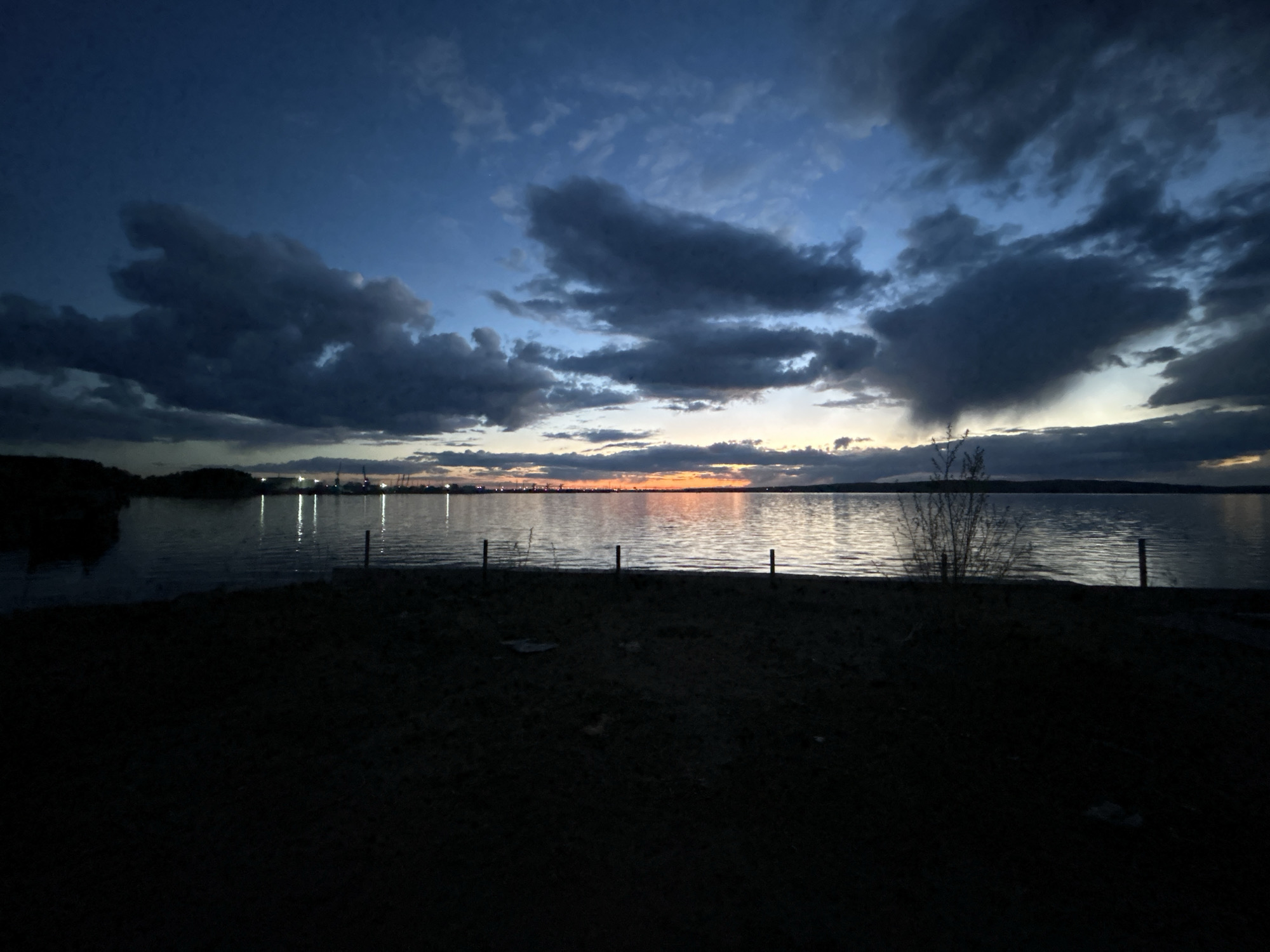 The image captures a twilight scene at a body of water, possibly in the city of Chaikovsky, Russia, based on the presence of industrial lights and the general landscape. Dark, heavy clouds dominate the sky, with the remnants of a sunset visible on the horizon. The water reflects the remaining light and the industrial lights in the distance. In the foreground, there is a dark, grassy area, obscuring the exact viewpoint. There are also posts along the shoreline and a small, bare tree or bush on the right.