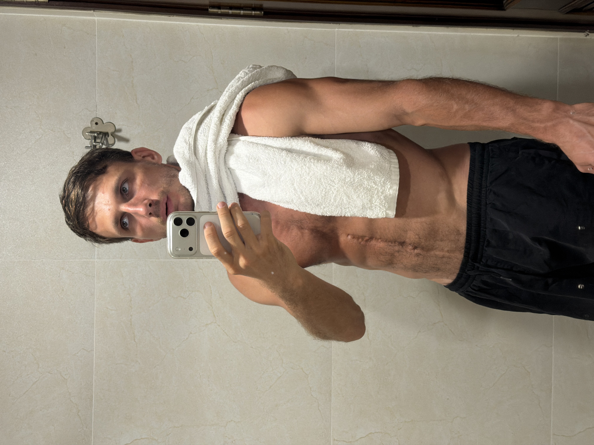 A man, appearing to be in his 20s or 30s with short dark hair and blue eyes, is taking a mirror selfie in what looks like a bathroom or changing room in Đà Nẵng, Vietnam. He is bare-chested, wearing black athletic shorts, and has a white towel draped over his left shoulder. He holds a smartphone with a clear case in his right hand, angled to capture his reflection. His expression is neutral to slightly surprised, with his mouth slightly ajar, as he looks towards the phone.
A distinctive, somewhat raised scar runs horizontally across the left side of his abdomen, extending from his central torso towards his flank. The background features light-colored, possibly off-white or beige, marble-patterned tiles on the wall. A small, metallic, cloud-shaped wall hook is visible on the tiled surface above his shoulder. A dark wooden or metal frame or ledge is visible at the top edge of the mirror's reflection. No discernible text is visible on the phone screen or elsewhere in the image. The lighting suggests an indoor setting, likely provided by artificial sources.