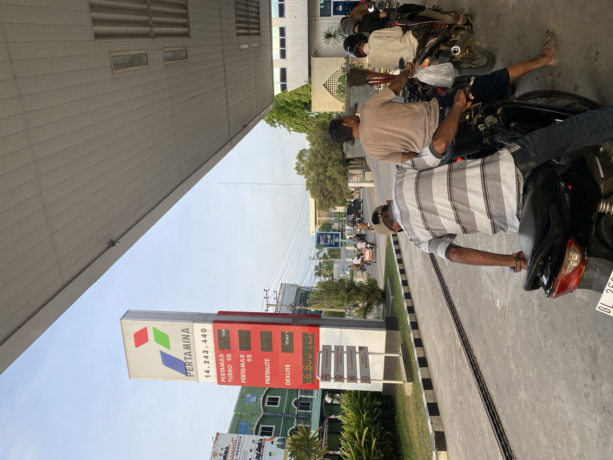 The image captures a scene at a Pertamina gas station in Lhokseumawe, Indonesia. A prominent Pertamina sign displays fuel prices. In the foreground, several people are waiting in line on motorcycles. The sky is visible with a bright, overcast appearance. The environment suggests a typical urban setting with buildings and vegetation in the background. The overall lighting indicates it is daytime.