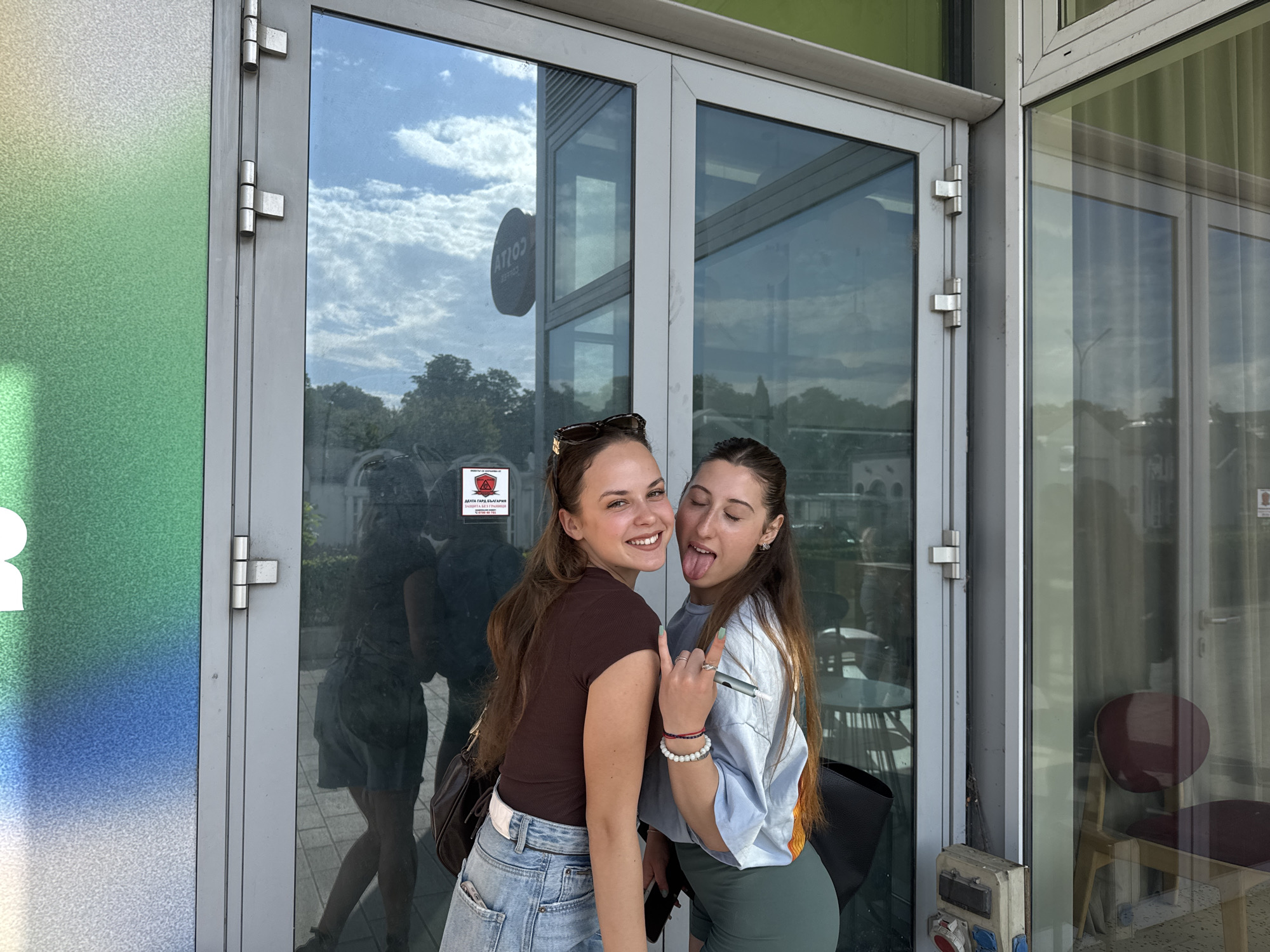 Two young adult females are posing in front of a glass building entrance in Unknown, Bulgaria. The left woman, with long brown hair, smiles directly at the camera. She wears a dark brown t-shirt, light blue ripped jeans, a white belt, and has sunglasses perched on her head. A dark shoulder bag hangs from her left shoulder. The right woman, also with long brown hair, sticks her tongue out and makes a "rock on" hand gesture with her right hand. She wears a light blue t-shirt, dark green cycling shorts, and has a black shoulder bag. Multiple bracelets (red, black, white) are visible on her right wrist, and she holds a slender, pen-like object.
Behind them, the glass double doors and adjacent windows reflect the outdoor environment, including a partly cloudy sky, green trees, and distant buildings. The reflection of a circular sign displaying "COTTO" (reversed) is visible in the upper left glass panel. On the lower left glass panel, a red and black warning sign with Cyrillic text, reading "ДАТА ГАРА БЪЛГАРИЯ" (DATA GUARD BULGARIA), is reflected. To the left of the doors, a wall panel features a vertical gradient transitioning from green at the top to blue and white at the bottom. Through the rightmost glass panel, the interior of the building is partially visible, showing a burgundy chair and a table. A grey electrical box with red and blue outlets is mounted on the lower right door frame.