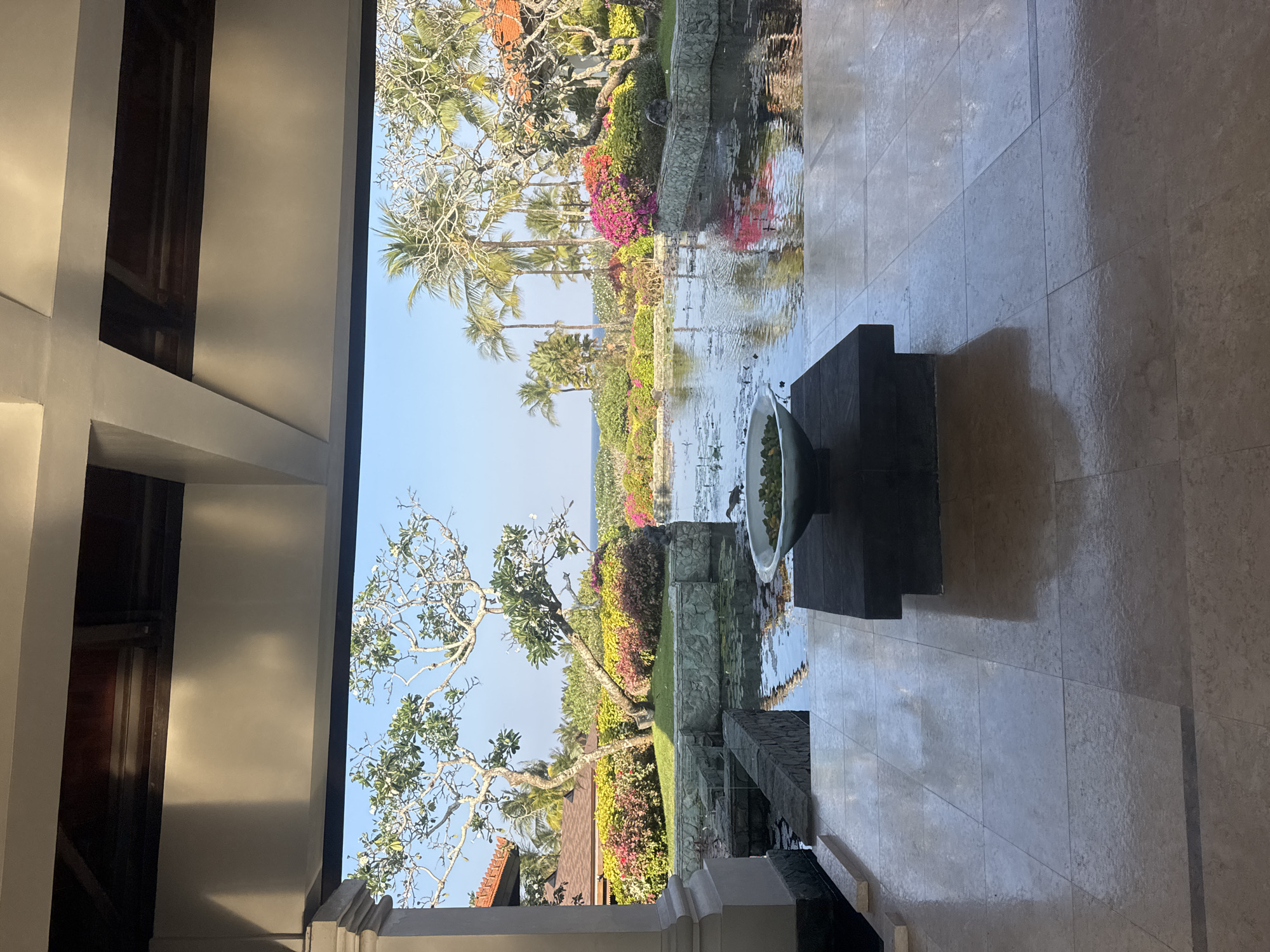This is a vertically oriented, eye-level, outdoor shot of a tranquil resort courtyard in Kuta Selatan, Indonesia, on a sunny day. The foreground shows a polished stone floor extending into a covered walkway. Beyond this, a serene water feature with lily pads and reflections of lush greenery dominates the scene. A stone-lined pond is filled with water, creating a reflective surface. In the middle of the pond, a large, shallow, light green bowl is filled with water and what appears to be green floating foliage. Stone walls surround parts of the pond, and a bird is visible on the far right edge of the pond.
Beyond the water feature, a vibrant garden bursts with colorful flowers in shades of pink, red, and yellow, interspersed with green bushes. Palm trees and other tropical trees with intricate branches are scattered throughout the landscape. In the distance, the bright blue of the ocean can be seen under a clear sky, with a hint of land or buildings on the horizon. The architecture of the resort is visible on the left and right sides, with tiled roofs and wooden beams supporting the covered walkway. The sunlight casts shadows on the floor and illuminates the tropical vegetation.