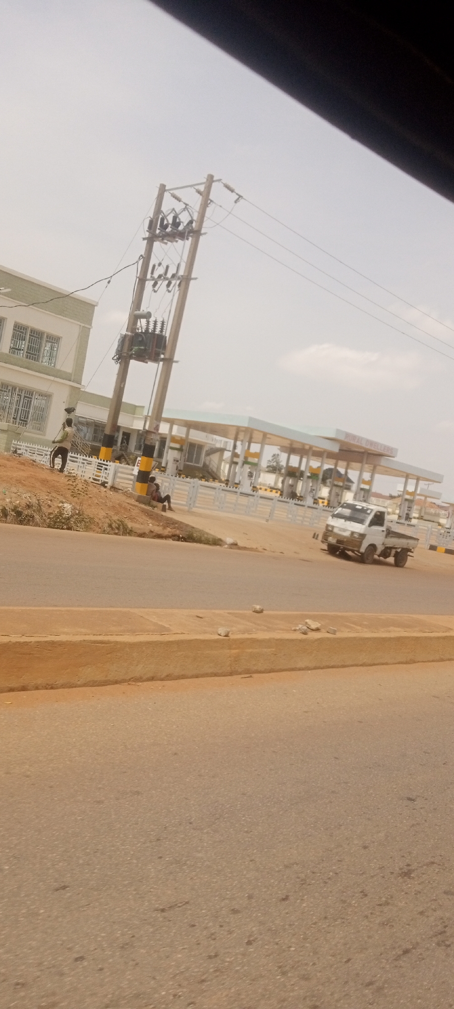 The image shows a roadside scene in Rido, Nigeria, featuring a building under construction next to an electrical power pole. Adjacent to the building is a gas station identified as "Kowac Dwellers." A small white truck is on the road. One person is standing near the building with a brush, and another individual is sitting at the base of the pole. The environment is hazy and sunlit.