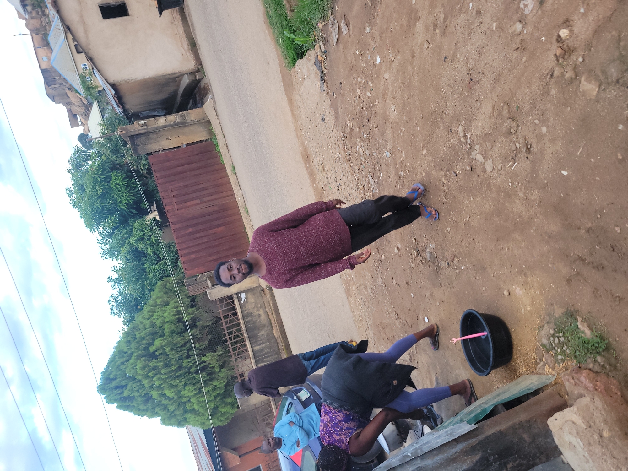 This image depicts an outdoor street scene in a residential area of Jos, Nigeria, during daylight hours under an overcast sky. The foreground consists of a dirt and gravel shoulder, transitioning to a paved road on the left and mid-ground.
Four individuals are present. On the far right, a young adult male with dark skin and a beard stands facing the camera, wearing a long-sleeved maroon sweater, dark trousers, and blue flip-flops. To his left, in the foreground, a woman with dark skin is bent over, partially obscured by a corrugated metal sheet. She wears a purple patterned top, a dark apron/skirt, and light blue leggings with dark flip-flops, attending to a black plastic basin containing a pink, hand-shaped object. Behind her, a dark blue sedan is parked on the road. Leaning against the car, an adult male with dark skin, wearing a light blue long-sleeved shirt and dark trousers, stands with his arms crossed, looking left. Another adult male, also with dark skin, stands further back between the woman and the car, facing partially away from the camera in a dark shirt and blue jeans.
The background features several residential buildings constructed into a hillside on the right, some with corrugated metal roofs and exposed rock formations. A reddish-brown metal gate and a concrete wall are visible behind the individuals on the road. A large, conical evergreen tree and other leafy trees are present behind the wall and gate. Utility wires traverse the sky. The ground exhibits scattered debris, small rocks, and sparse patches of green vegetation.