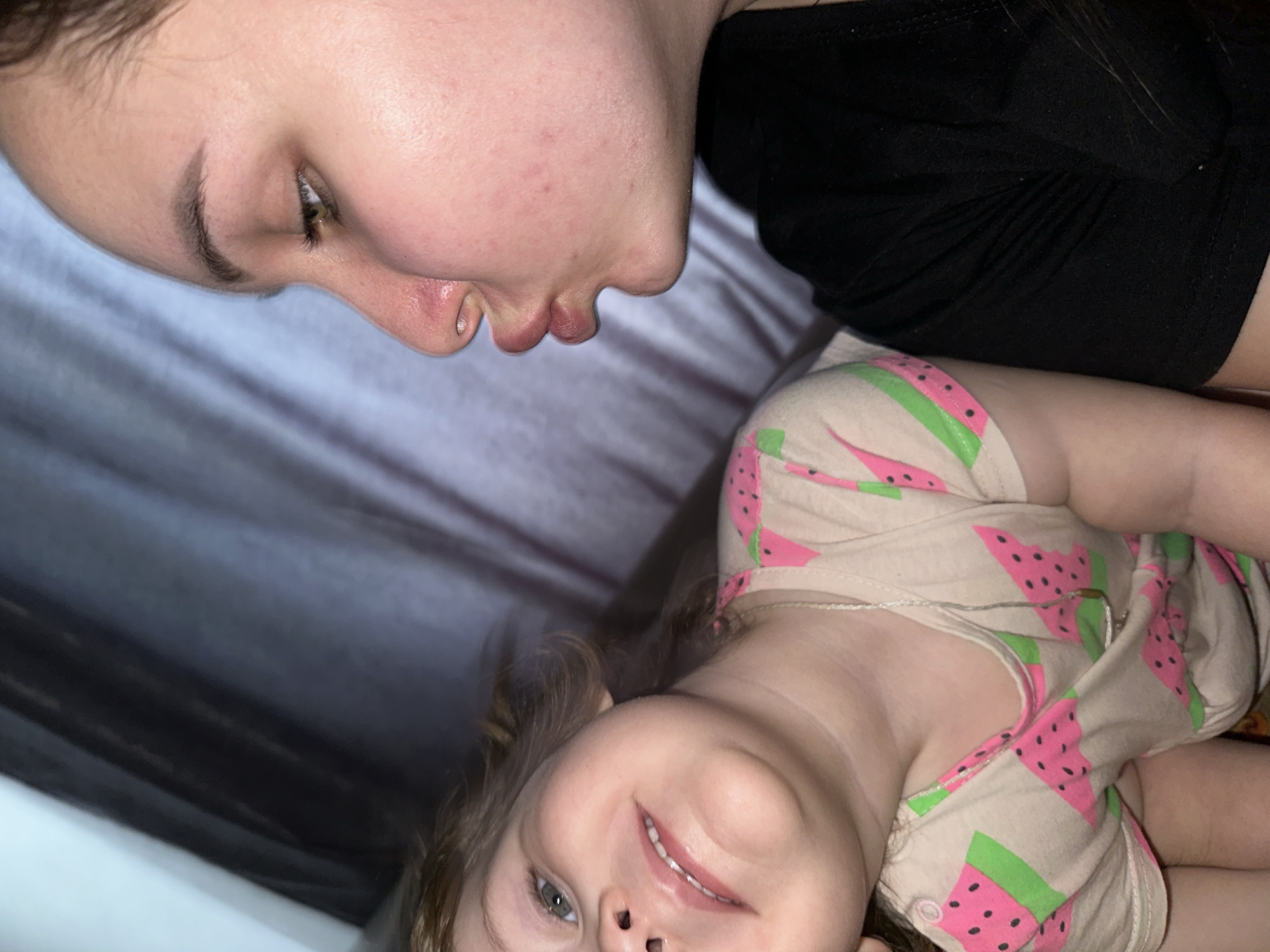 The image shows two people, likely a mother and child, lying down indoors. The adult, with fair skin and dark hair, is wearing a black shirt. The child, also fair-skinned with light brown hair, is wearing a light-colored shirt with a pink watermelon pattern. They appear to be on a bed with a gray patterned sheet. The location is not identifiable from this image as being in the city of Chaikovsky, Russia or any other specific location.