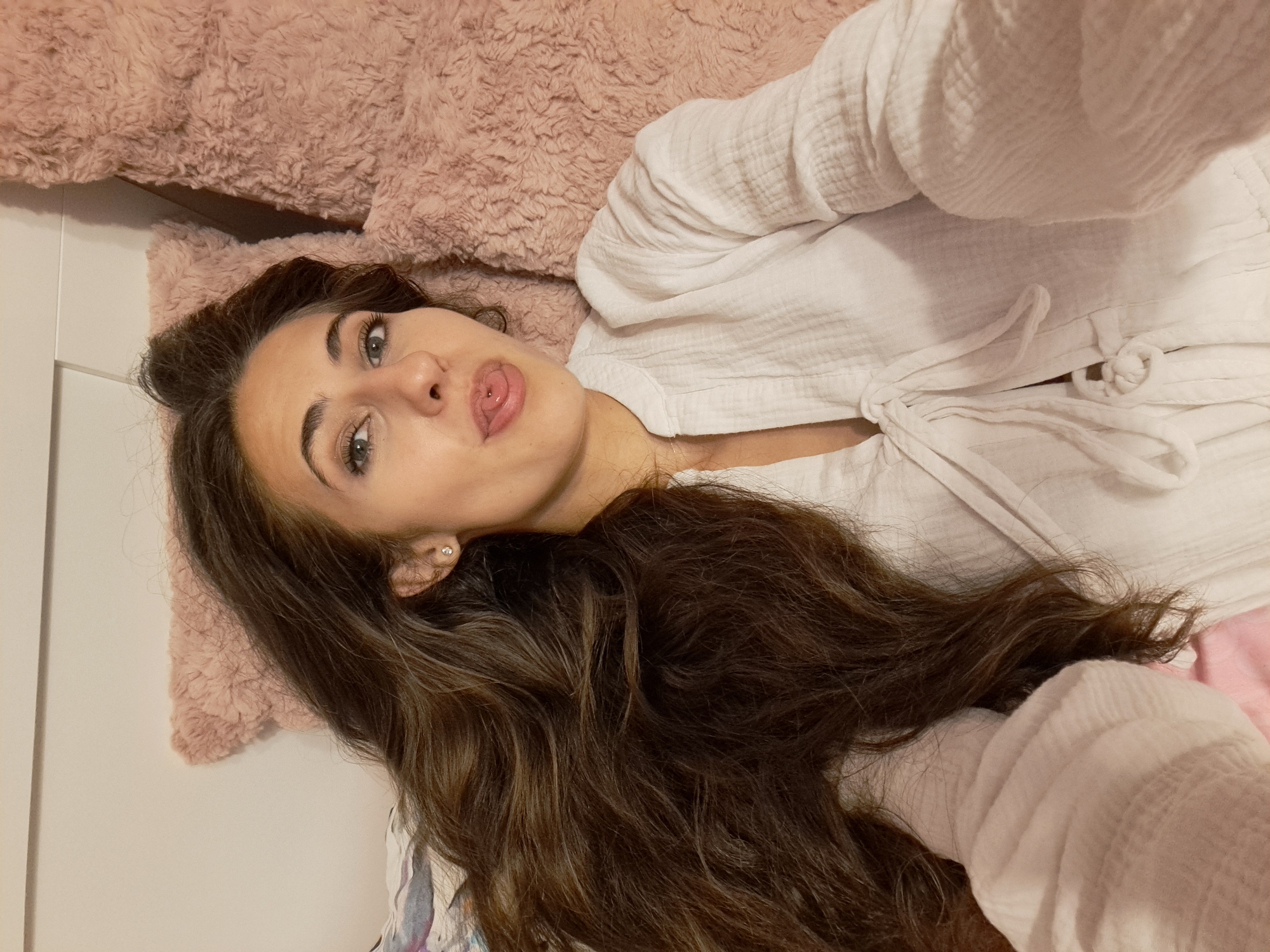 A young woman with long, dark, wavy hair and light-colored eyes is lying on a pink, fuzzy surface, possibly a blanket or pillow. She is wearing a white, long-sleeved top with a textured fabric and ties at the front. Her head is resting on a smaller, similar pink, fuzzy pillow. She is looking directly at the camera with a playful expression, sticking her tongue out slightly. Her left ear is visible, adorned with a small, sparkly stud earring. The lighting appears to be soft and even, suggesting an indoor setting. Part of a white surface, possibly a wall or headboard, is visible behind her. The overall mood is relaxed and informal.