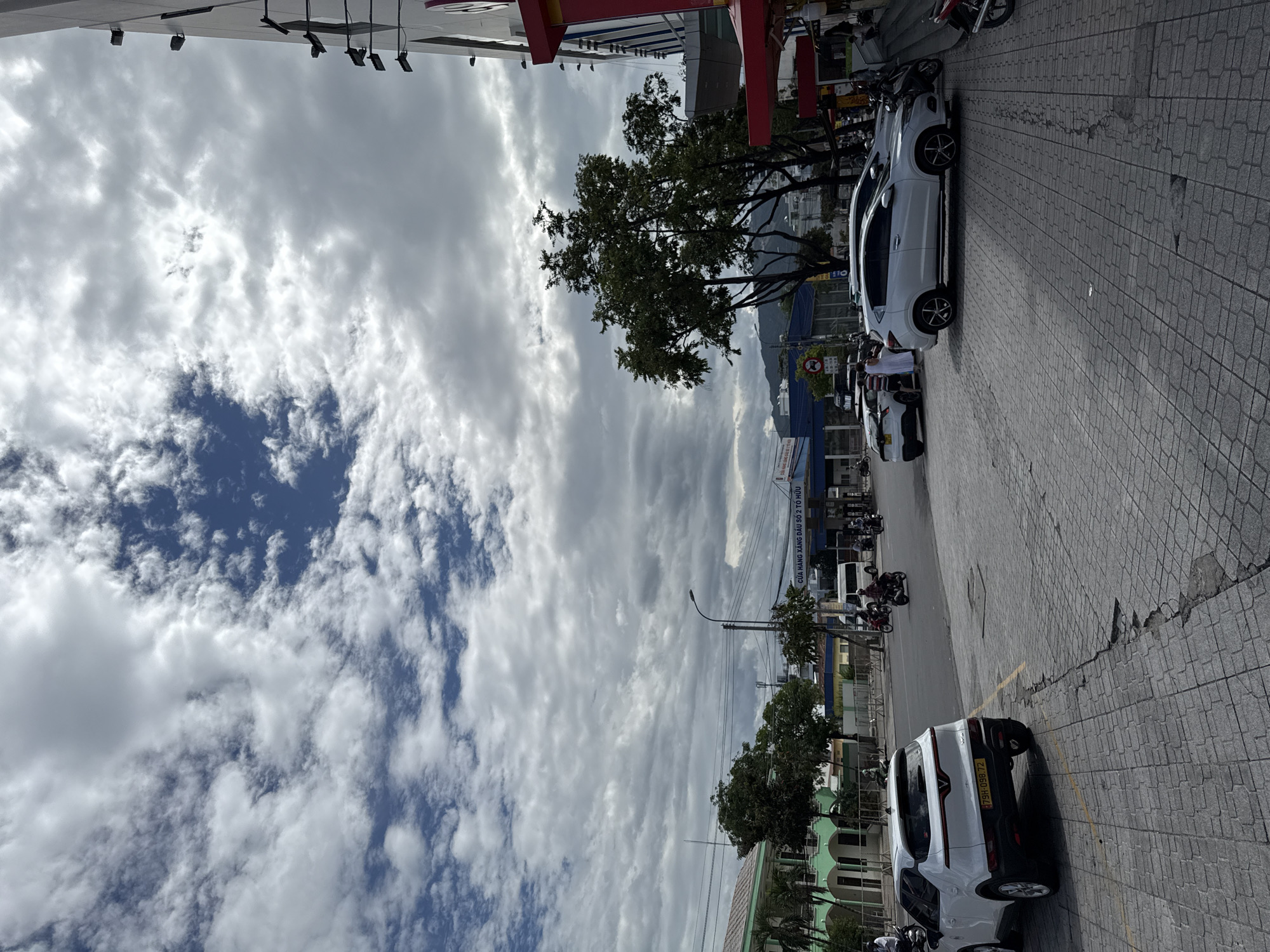 The image depicts a street scene in Nha Trang, Vietnam. The sky is filled with scattered clouds. In the foreground, a white SUV is parked on a paved walkway. Further down the street, other vehicles, including motorcycles and a white van, are visible. Buildings line the street, some with green facades and arched windows. Trees are planted along the roadside. A sign with Vietnamese text is visible on a structure that appears to be a gas station. Several individuals are seen on the street and sidewalk, some riding motorcycles. The overall scene suggests a commercial area in a Vietnamese city.