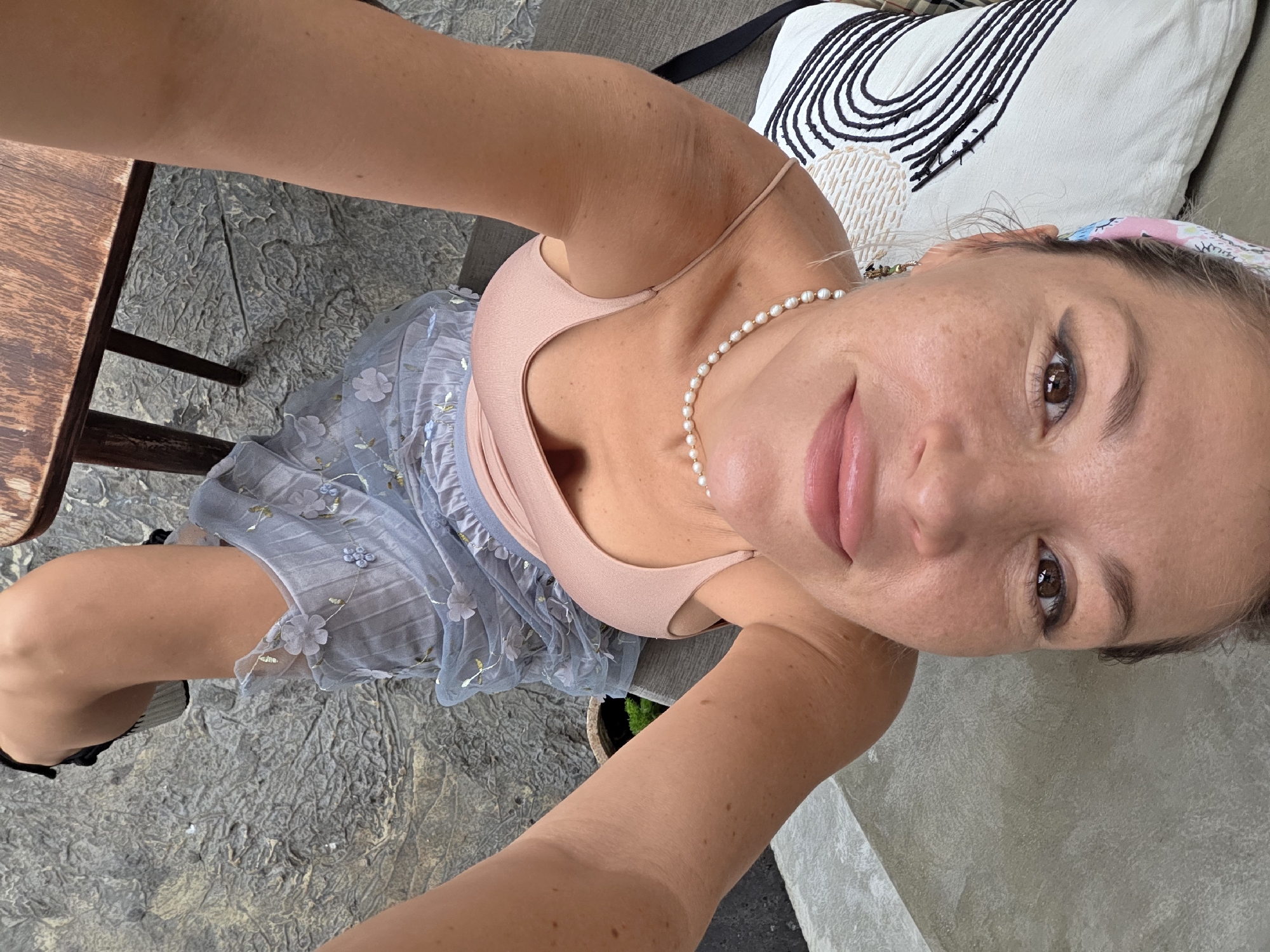 A woman is captured in a high-angle selfie taken outdoors. She is seated, with her legs crossed, wearing a light pink tank top and a sheer, pale blue skirt adorned with delicate floral embroidery. A single strand of pearls rests on her décolletage. The background reveals a textured, greyish ground, possibly concrete or stone, with a hint of a wooden table and chair on the left. Behind her, a textured cushion with black and white abstract patterns is visible. The scene suggests a casual, relaxed setting, possibly a cafe or patio in North Kuta, Indonesia. The lighting indicates it is daytime, with natural light illuminating the scene. The woman's expression is bright and engaging, with a slight smile and direct eye contact with the camera, conveying a positive and friendly emotion. There is no visible text in the image.