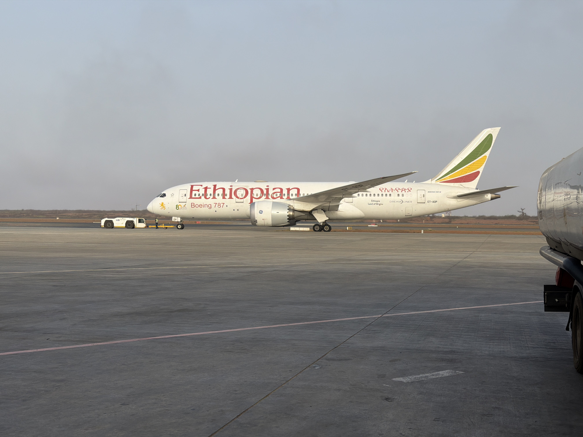 The image depicts an Ethiopian Airlines Boeing 787 Dreamliner on an airport tarmac. The aircraft is painted white with the word "Ethiopian" in red along the fuselage. The tail fin has the colors green, yellow, and red. The registration number "ET-AOP" is visible near the tail. In the background, there is a small vehicle, and to the right, part of a fuel truck is visible. The sky is overcast, suggesting hazy conditions. The scene appears to be on the ground, as there are ground crew vehicles visible. The image was taken in the city of Communauté rurale de Diass, Senegal.