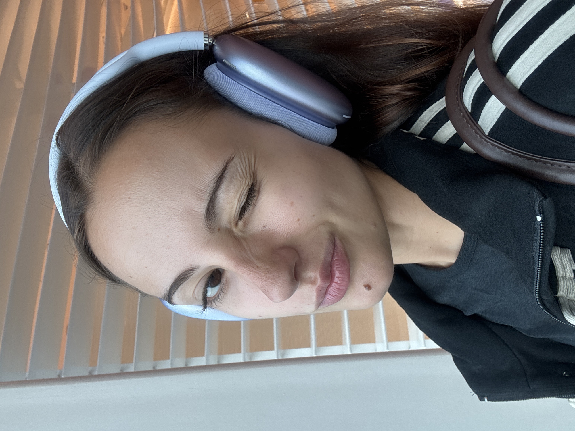 A woman with brown hair is wearing white and light blue headphones on her head. She is wearing a dark-colored hooded jacket, a striped shirt, and a black t-shirt underneath the jacket. The woman is looking at the camera with one eye partially closed. The background shows what appears to be a window with light beige-colored blinds. The location could be Singapore, Singapore, however, there is no visible evidence to indicate where this image was taken.
