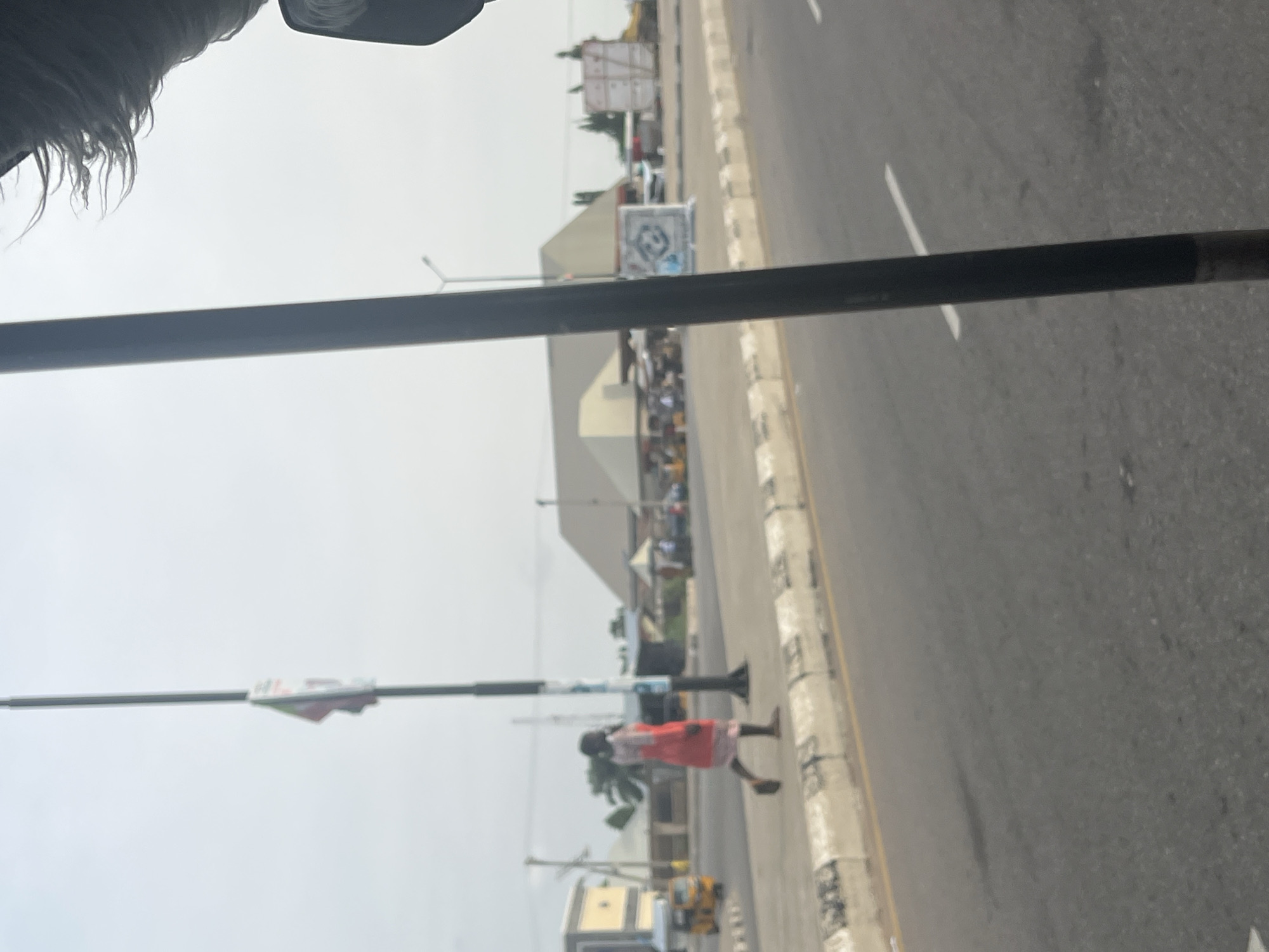 The image depicts a street scene in Jalingo, Nigeria. A person wearing a red and white patterned dress is walking on the sidewalk adjacent to a road. The sidewalk is bordered by a concrete curb. In the background, there are buildings, some with awnings, and a yellow tricycle. A tall pole with a flag is visible. The sky is overcast. The perspective suggests the photo was taken from a moving vehicle.