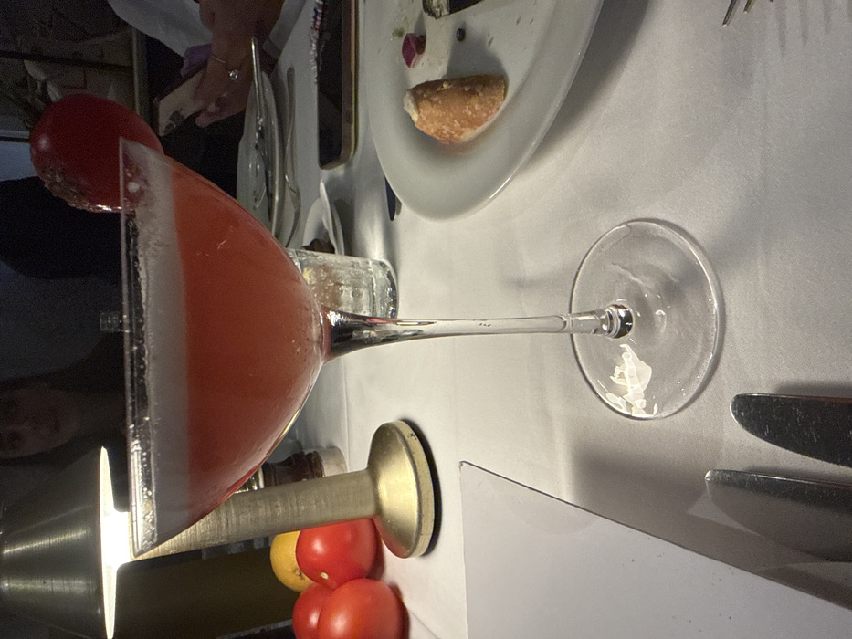 A red cocktail in a martini glass sits on a white tablecloth. The rim is frosted with white powder. Garnished with a cherry, the glass is supported by a brass stand. A plate with leftover food and silverware are present. Three tomatoes and a lemon are visible next to the glass stand. A person's hand is seen reaching for the plate.