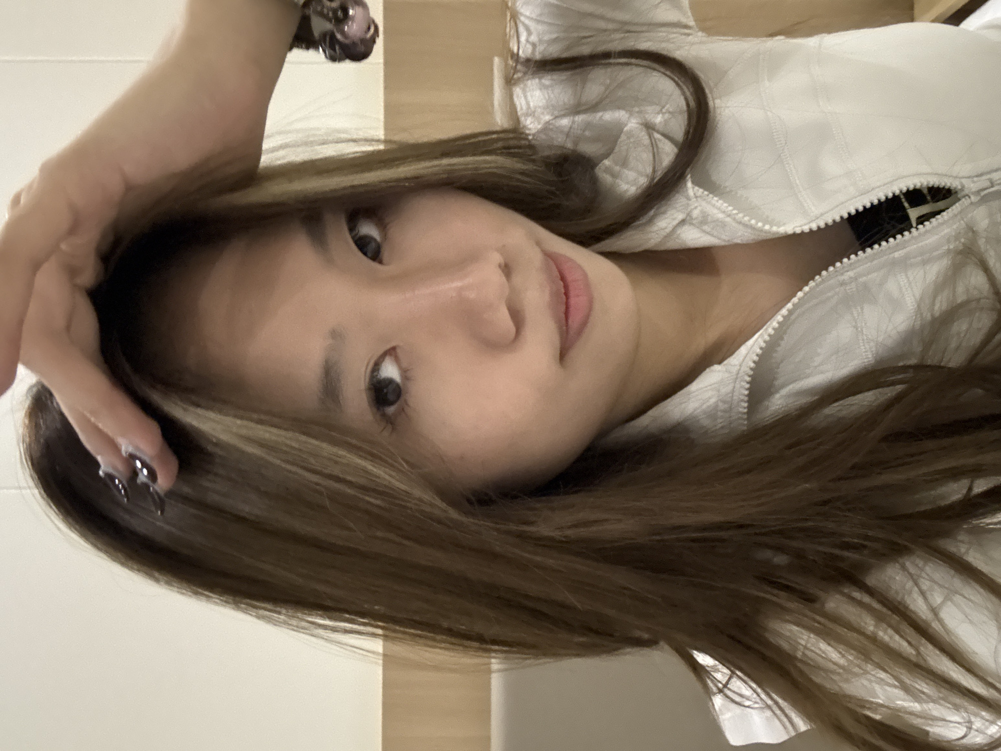 A close-up, rotated portrait of a young Asian woman with long, light brown hair and highlighted streaks. She is wearing a white quilted jacket with the zipper partially open. Her left hand is raised near her head, displaying dark-colored nail polish. The background is a blurred combination of white and beige tones, suggesting an indoor setting. There is no indication that this photo was taken in Dunhuang City, China.