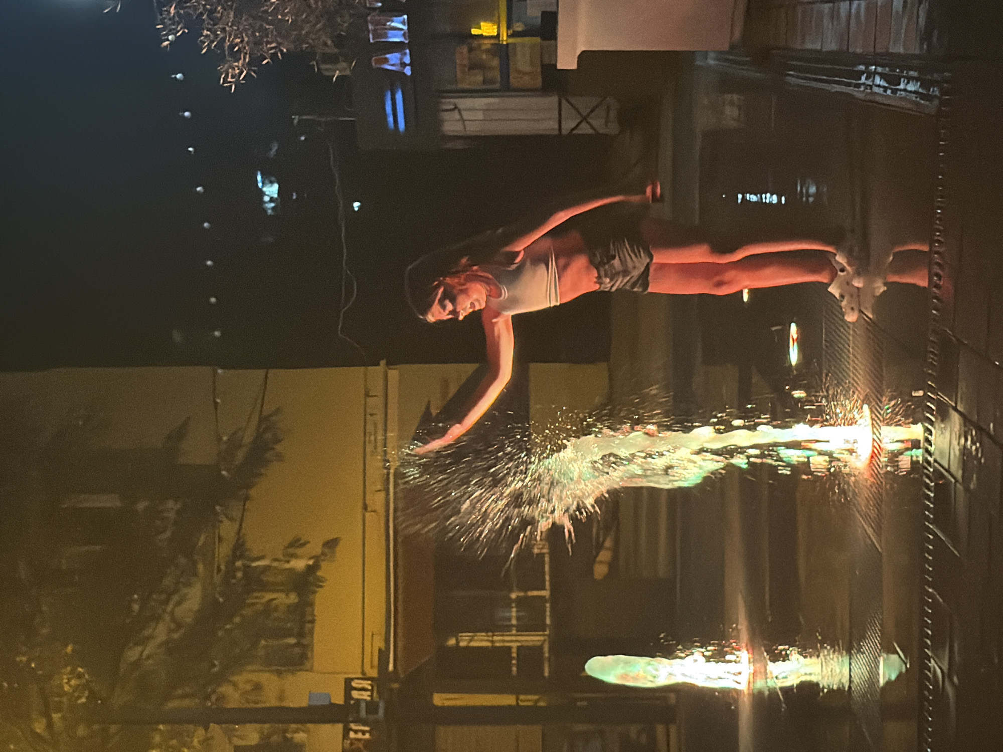 The image depicts a person performing a water-based act, likely a dance or acrobatic stunt, at night. The subject is a young woman with long hair, wearing a light-colored tank top and dark shorts. She is positioned horizontally, with her body extended as if diving or jumping. Her arms are outstretched, and she is creating a large splash of water that is illuminated by colorful lights, possibly from stage lighting or reflections.
The setting appears to be an outdoor venue, possibly a performance space or a public area with a water feature. The background is dark, suggesting it is nighttime. There are hints of buildings and structures in the distance, with some lights visible. Trees with sparse foliage are also present in the scene. The surface beneath the water is reflective, possibly a pool or a wet floor, and it also reflects the colorful lights, creating a vibrant and dynamic effect.
The activity captured is a moment of action, where the subject is interacting dynamically with water. The spray from the water is frozen in motion, highlighting the intensity of the movement. The lighting creates a dramatic and somewhat surreal atmosphere, with the colorful glow enhancing the visual appeal of the water splash and the performer. No specific emotions are clearly discernible from the subject's face due to the distance and lighting, but the overall impression is one of athletic performance and artistic expression.
There is some faint, possibly Cyrillic text visible on a sign in the lower left portion of the image, but it is too blurry to be read clearly. The location context is Partenit, Ukraine.