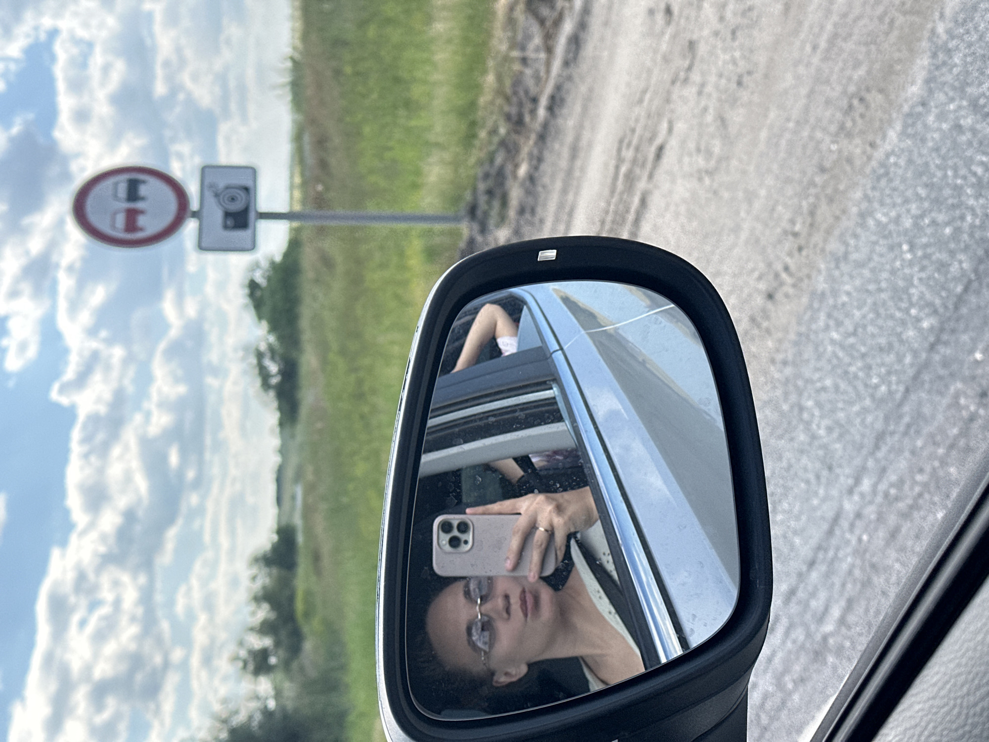 In the foreground, a reflection of a young woman is visible in the side-view mirror of a car. She is wearing glasses and taking a selfie with her phone. Her lips are pursed in a "duck face" pose. Behind her, another person's arm is visible. The car appears to be on a dirt road, with grassy fields and trees in the background. A road sign indicating "no overtaking" and a camera symbol is visible in the distance, possibly in the city of Ferma Sovkhoz Balykleyskiy, Russia. The sky is cloudy with patches of blue.