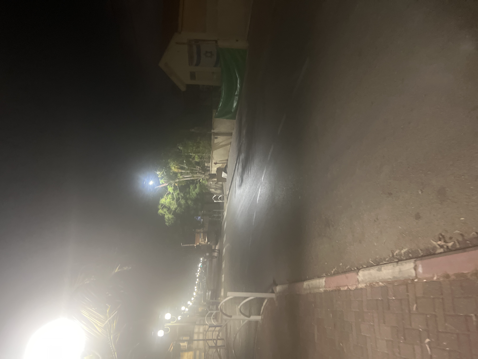 The image depicts a nighttime scene on a street in Or Akiva, Israel. The street is paved and appears to be a thoroughfare with multiple lanes. On the left side of the street, there is a sidewalk made of red brick pavers, bordered by a concrete curb with red and white sections. Several streetlights with white globes illuminate the area, casting a bright glow.
Further down the street, the lights of passing vehicles are visible, indicating traffic. Two figures are walking across the street in the mid-ground. On the right side of the street, a low cream-colored building is visible, with a large green banner hung on its wall. Above the banner, an Israeli flag is displayed. Trees and other vegetation are present along the right side of the street, some illuminated by the streetlights. The overall environment suggests a somewhat suburban or town setting at night, with artificial lighting dominating the scene.