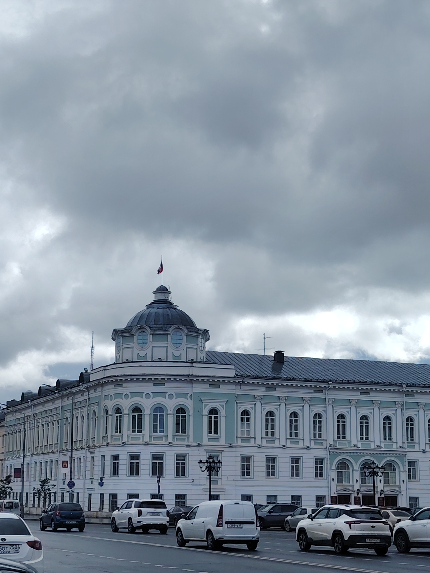 The image depicts a wide street in Tver, Russia, with a prominent, ornate, light blue and white building occupying the right side of the frame. The building features multiple windows, arched entrances, and a distinctive dome topped with a Russian flag. The sky is heavily overcast with thick, grey clouds. Several cars are visible on the asphalt road, including a white SUV, a dark blue hatchback, and a white van. The architecture suggests a historical or governmental structure within the city.