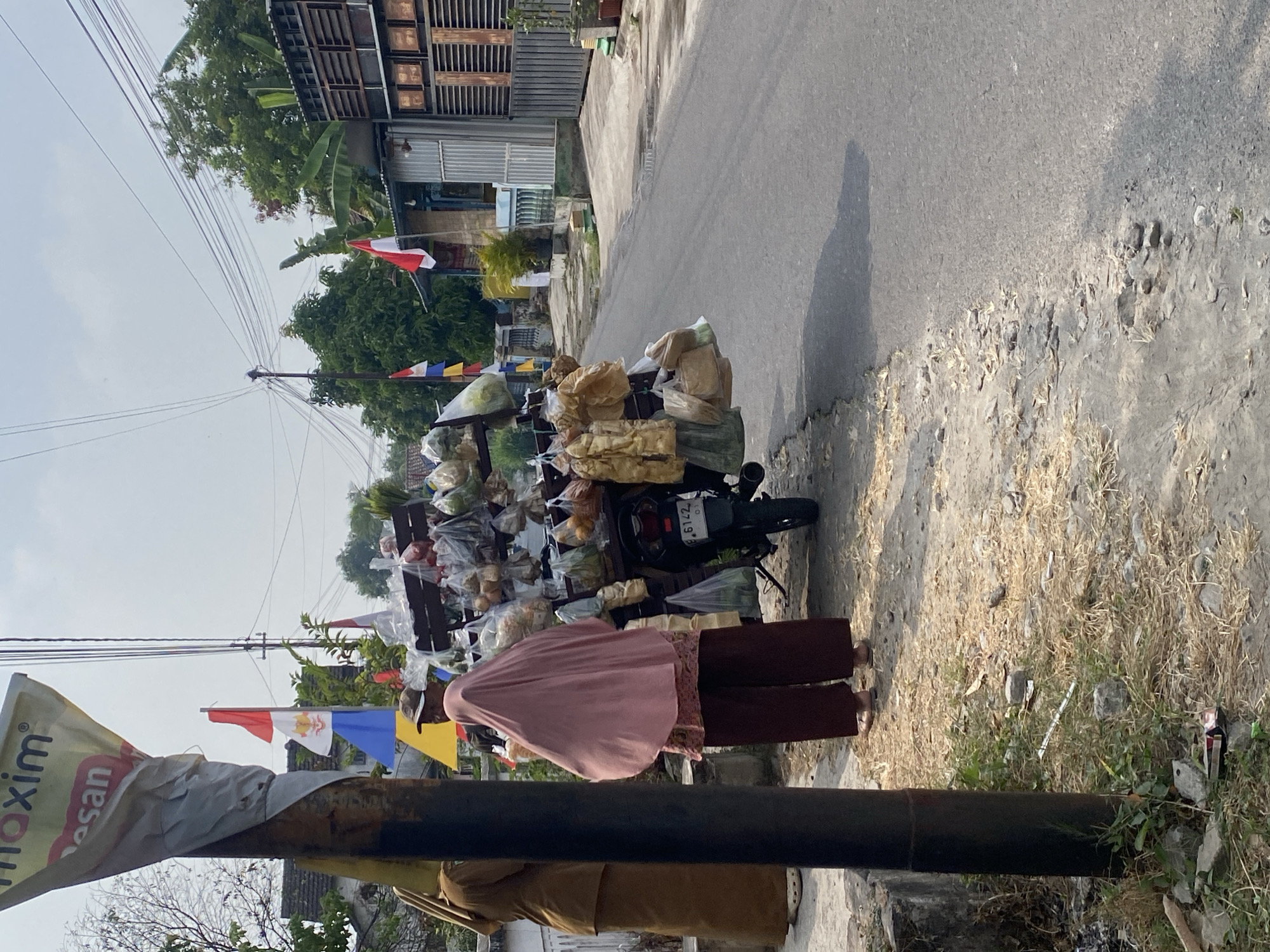 An outdoor daytime medium shot captures a street scene in Kraton, Indonesia. A motorcycle-pulled cart is laden with various bagged food items, suspended from a rack. Two individuals are present: one in the foreground wearing a pink headscarf and maroon pants, facing away from the camera. To the left and slightly behind, another person stands dressed in a brown uniform. In the background, a street is visible with houses lining one side. Several Indonesian flags are displayed, some attached to poles near the cart. The ground is paved with asphalt on the right and a dirt path with dry grass and scattered debris on the left. A utility pole with electrical wires runs across the upper portion of the image, and a sign with text partially obscured is attached to a pole in the foreground.