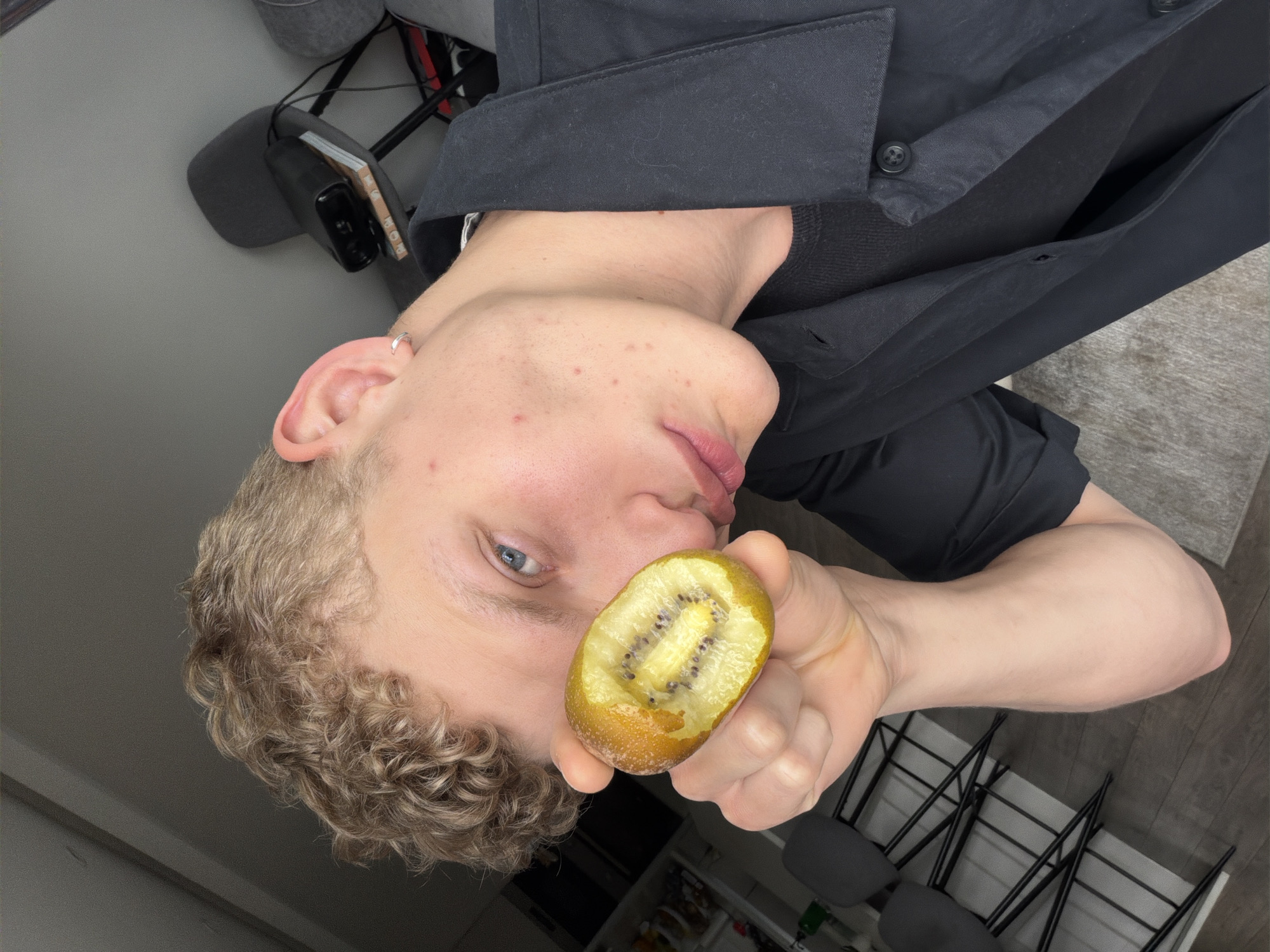 A close-up, selfie-style portrait of a young man with curly blonde hair and blue eyes. He is holding a halved kiwi fruit up to his right eye, partially obscuring his face. The man is wearing a black collared shirt over a black t-shirt. His skin has visible blemishes. In the background, out of focus, are chairs and what appears to be a shelf with items. The overall lighting suggests an indoor setting, possibly a home. There is no discernible information to place this image in London, United Kingdom.