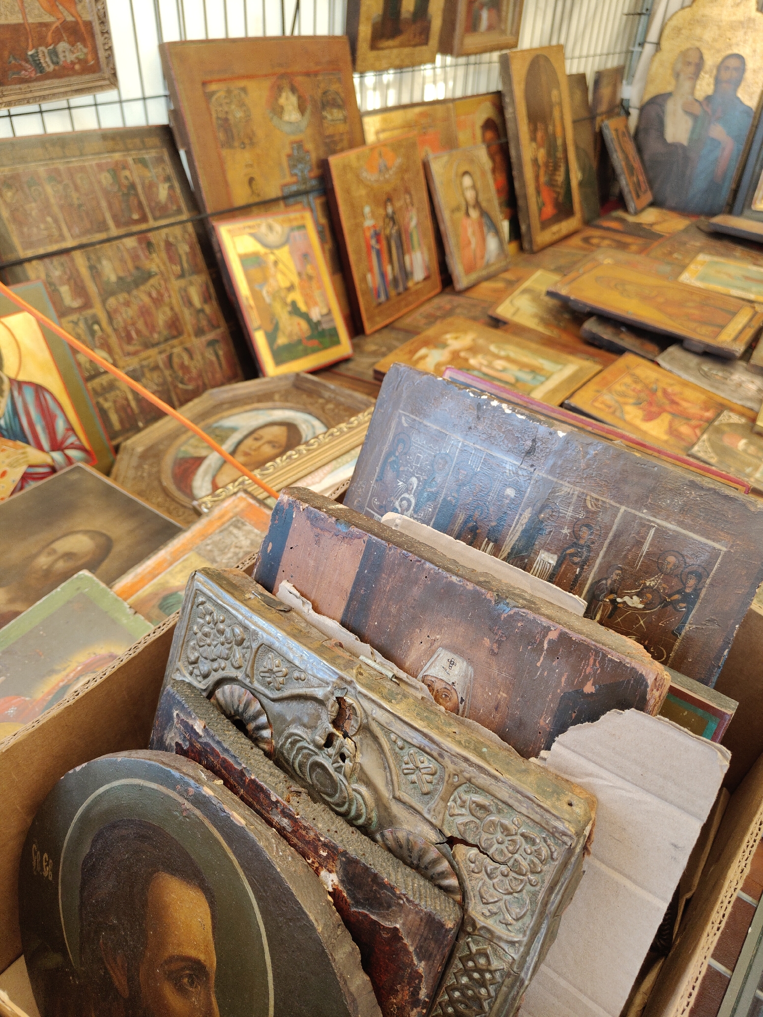 The image shows a close-up view of numerous religious icons, primarily Eastern Orthodox, displayed on shelves and in boxes. The icons vary in size and condition, with some appearing antique and worn, while others seem newer. The subjects depicted are religious figures, saints, and scenes from the Bible, rendered in a traditional style with gold leaf backgrounds and vibrant colors.
The icons are densely packed together, suggesting a market stall or a religious artifact shop. The arrangement is somewhat chaotic, with some icons leaning against each other and others stacked horizontally. The lighting is bright, casting shadows that emphasize the textures and details of the icons. The overall impression is one of a rich collection of religious art, possibly for sale or display.
The provided context indicates that this scene is located in Gdańsk, Poland. However, the image itself does not contain any direct visual cues that would allow for a specific determination of its location within the city. The background is blurred and does not provide architectural or geographical context.