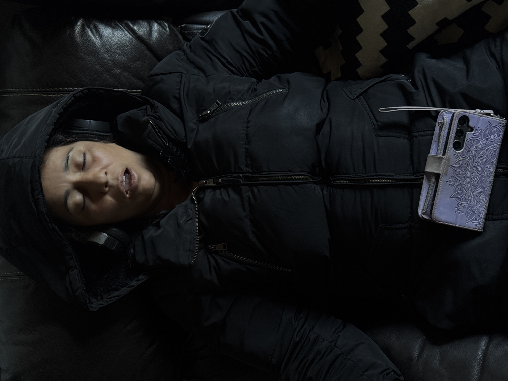 An individual is depicted lying supine on a dark leather sofa in an indoor setting within Esch-sur-Alzette, Luxembourg. The person, an adult female with dark hair, has her eyes closed and mouth slightly ajar, suggesting sleep or unconsciousness. She is wearing a dark, possibly black, hooded puffer jacket, with the hood pulled up over her head. Dark over-ear headphones are visible beneath the hood. A lavender-colored smartphone wallet case, featuring an embossed mandala pattern and a visible smartphone with three camera lenses, rests on her lower abdomen. A light-colored wrist strap extends from the case. A patterned pillow, displaying a black and white chevron design, is partially visible to the right of the individual. The scene is dimly lit, with shadows prominent.