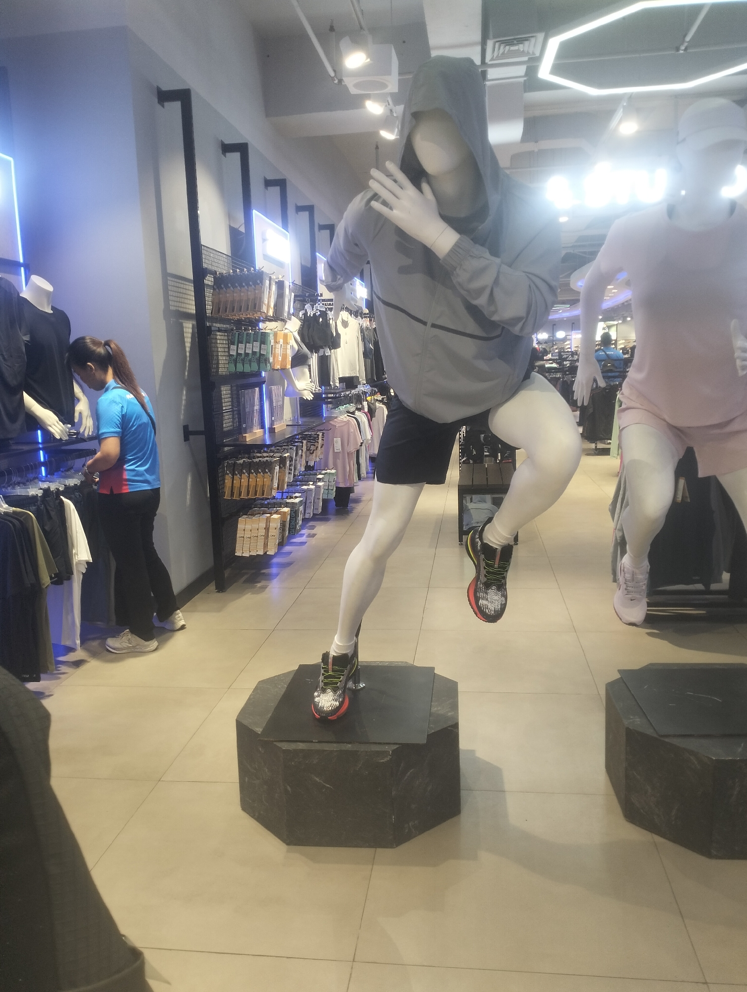 This image depicts the interior of a retail store, likely a sportswear shop, in Surabaya, Indonesia. The scene features two mannequins positioned as if running. The mannequin on the left is wearing a grey hooded jacket and black shorts, with one leg raised in a mid-stride pose. It is standing on a dark hexagonal pedestal. The mannequin on the right is wearing a pink t-shirt and white leggings, also in a running posture.
In the background, clothing racks display various apparel, including t-shirts and what appear to be athletic shorts. A woman wearing a blue and red shirt and black pants is visible in the mid-ground, interacting with the merchandise on a rack. The store has a tiled floor and fluorescent lighting overhead, with some hexagonal light fixtures. The overall environment suggests a commercial space focused on sports clothing.