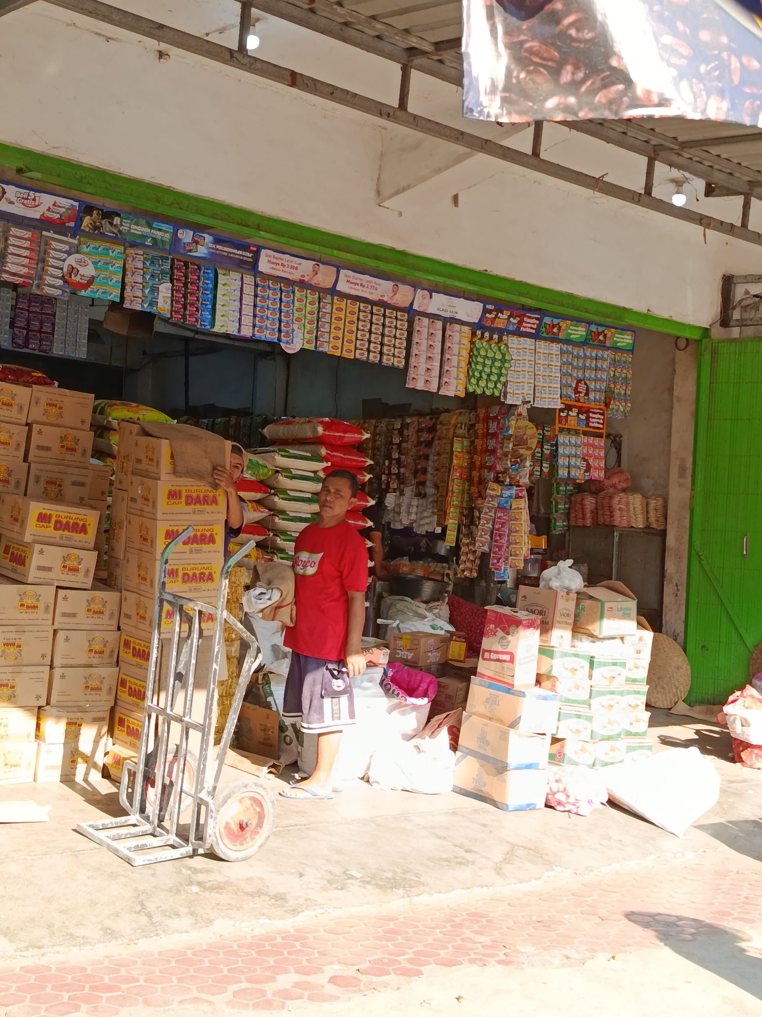 A man in a red t-shirt and blue shorts stands on a paved walkway in front of a storefront in Blindungan, Indonesia. He is holding a hand truck with several boxes stacked on it. The storefront is filled with various products, including bags of rice and boxed goods. The man appears to be in the process of moving or organizing inventory. The sun is shining, casting shadows on the ground.