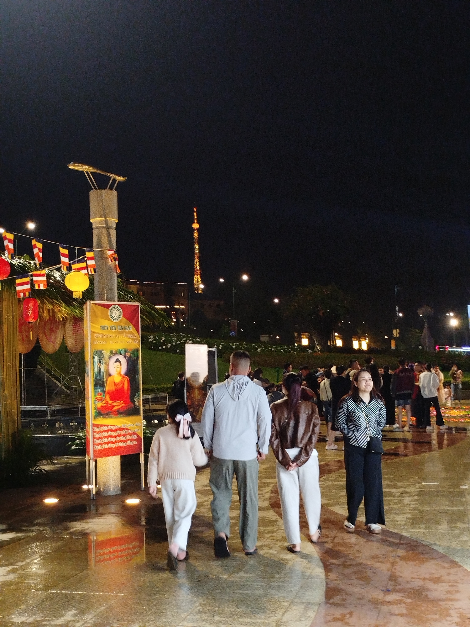 People are walking through a public space at night, in Da Lat, Vietnam. A family of four is walking towards a crowd of people in the background. To the left of the family is a display with the image of a sitting Buddha on a sign. There are lanterns and Buddhist flags strung above the display. The ground is wet and reflecting light. A tall communications tower is visible in the background.